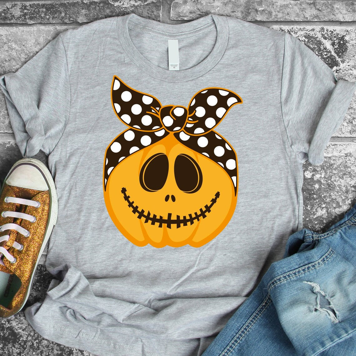 The Smiling Pumpkin Halloween T-shirt is a premium quality Bella Canvas gray t-shirt featuring a smiling jack-o'-lantern adorned with a black and white polka dot headscarf. It’s styled alongside one glittery sneaker and a pair of rolled-up jeans on a stone backdrop, offering ultimate comfort with its soft textile flex print.