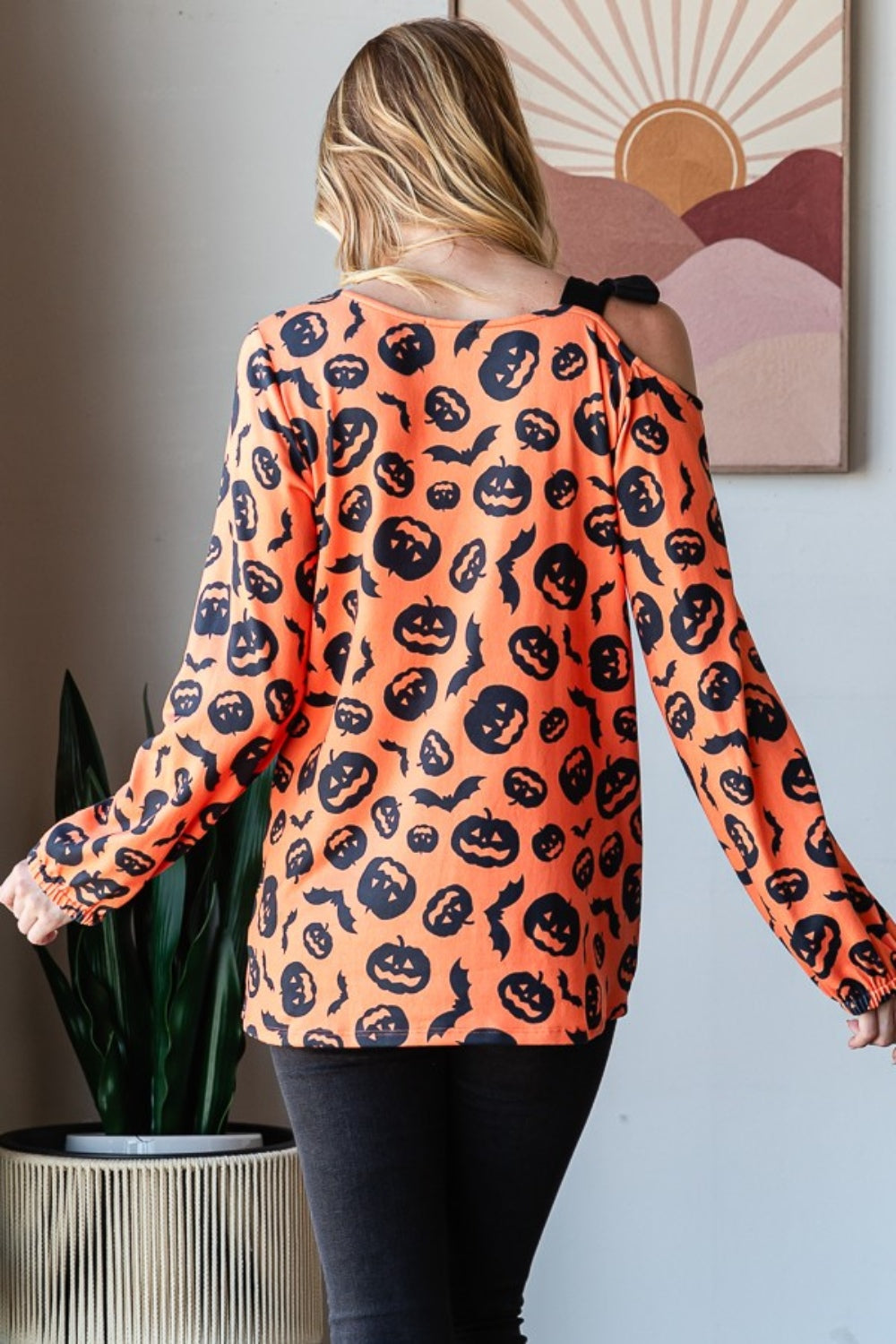 A woman smiles while modeling the Heimish Pumpkin Print One Shoulder T-Shirt, featuring an off-shoulder, long-sleeve design that is perfect for fall season fashion. She stands indoors with a plant and wall art in the background.