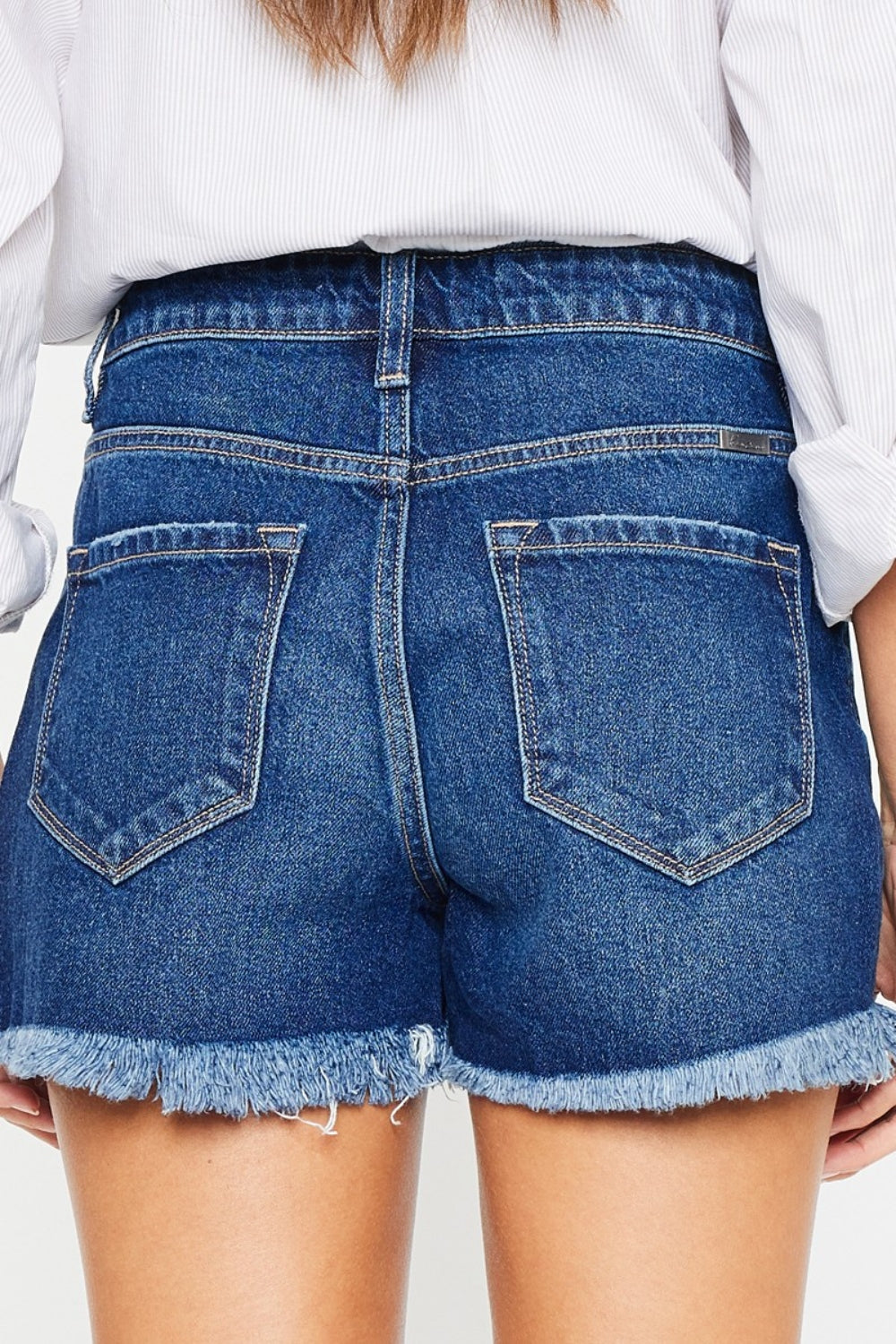 A woman with long dark hair wears a white blouse tied at the waist over a white top and Kancan High Rise Frayed Hem Denim Shorts, posing with one hand in her pocket against a plain background, embodying the perfect summer wardrobe.