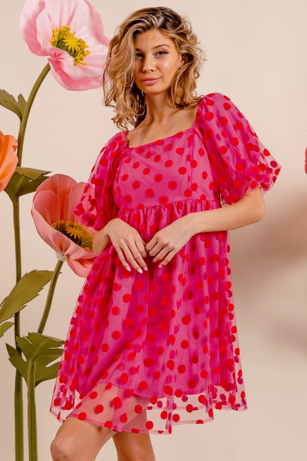 Wearing the BiBi Polka Dot Mesh Puff Sleeve Dress, a woman poses gracefully with one hand on her head. The vintage-inspired pink dress adorned with red polka dots enhances her elegance as she stands amidst large artificial flowers.