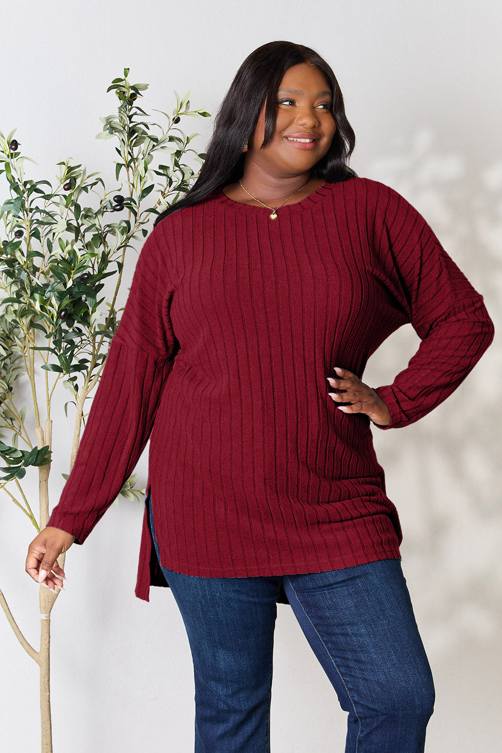 A person with shoulder-length hair smiles while wearing a beige ribbed sweater called the Basic Bae Full Size Ribbed Round Neck Long Sleeve Slit Top, along with matching beige pants, showcasing a basic style. They stand next to a green plant.