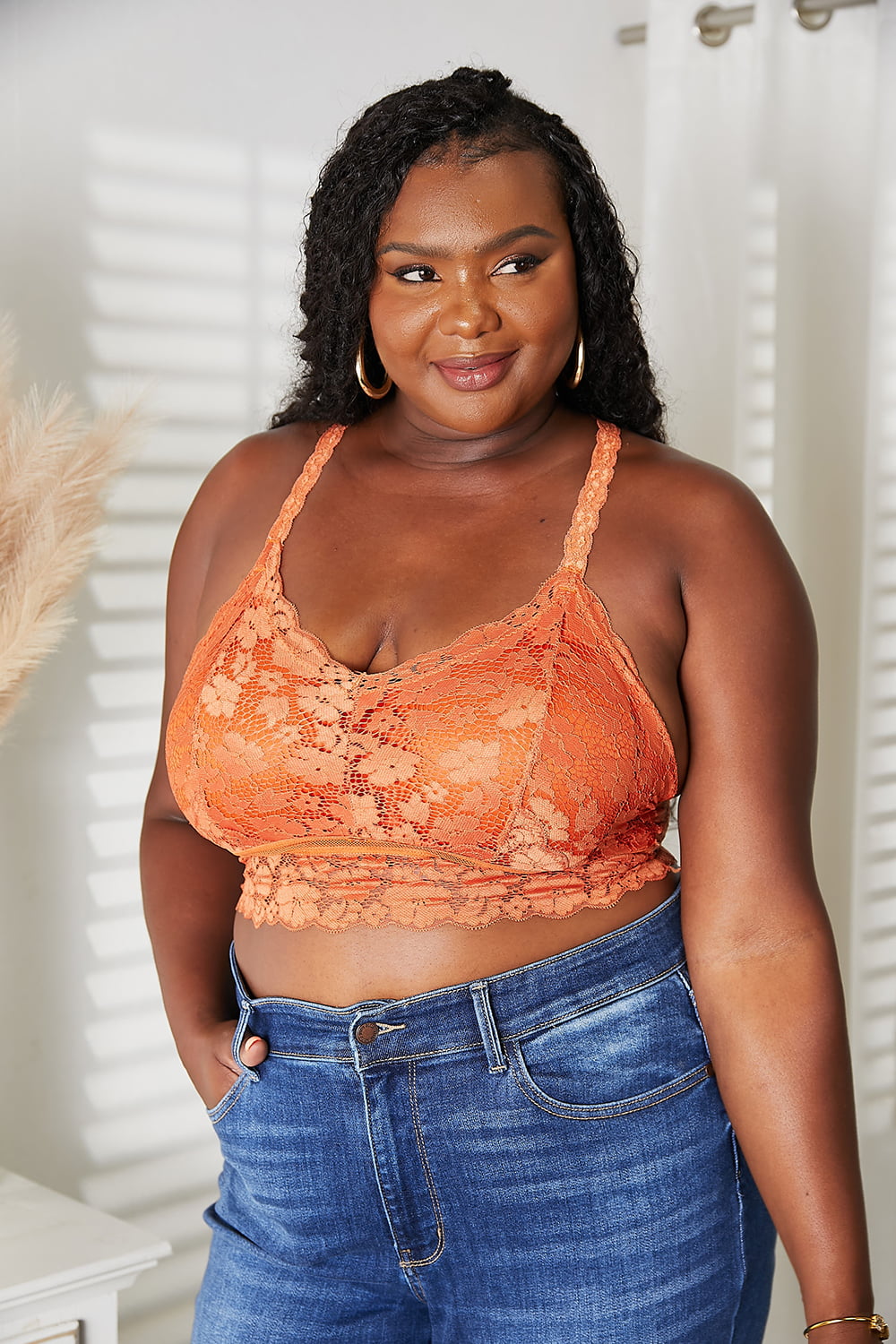 A woman with long curly hair wears the JadyK Juliette Full Size Crisscross Lace Bralette paired with blue jeans, standing beside a white vase with dried plants.