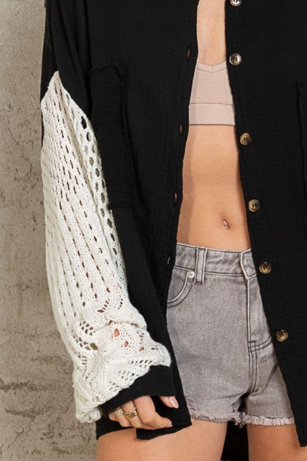 A woman with blonde hair stands against a gray textured wall, wearing the POL Openwork Lantern Sleeve Button Down Gauze Shirt—an elegant, lightweight, and breathable black button-up shirt with white knit sleeves. She pairs it with a beige top and gray shorts, creating a chic ensemble.