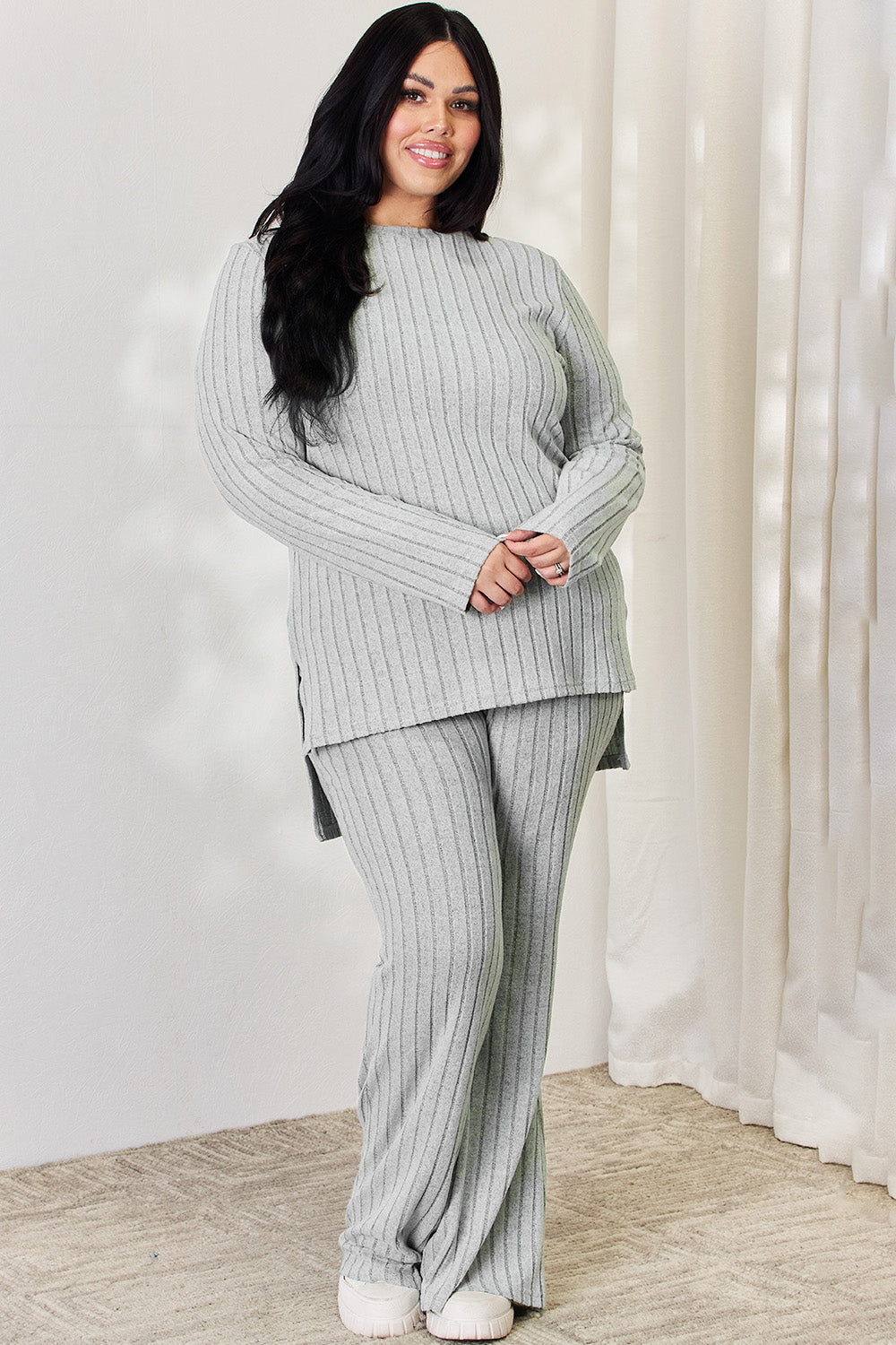 A person wearing the Basic Bae Full Size Ribbed High-Low Top and Wide Leg Pants Set in light gray, made from stretchy material, stands indoors in front of a white curtain.