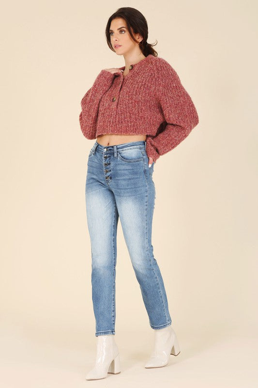 A person is wearing the Melange multicolor sweater top, characterized by textured pink fabric and featuring raglan sleeves along with large buttons, paired with light blue jeans, standing against a plain background.