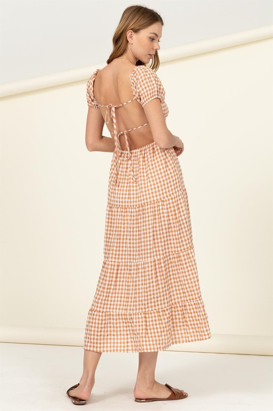 Wearing the "Somewhere to Go Tie-Back Gingham Print Maxi Dress," which features a black and white gingham print with puffed sleeves, a tiered skirt, and elegant tie-back detail, a person poses stylishly paired with black sandals.