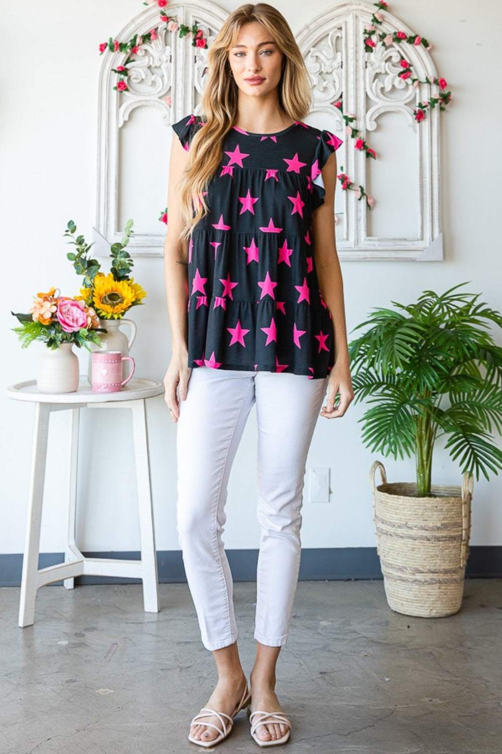 The woman stands elegantly in front of a floral arch, wearing the Heimish Full Size Round Neck Star Tiered Top paired with white pants. The round neck design of her top features a captivating star pattern, perfectly complementing the vibrant blooms behind her.