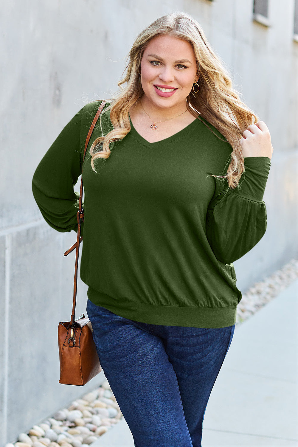 A woman with brown hair is standing against a concrete wall, wearing the Basic Bae Full Size V-Neck Lantern Sleeve Top in teal and blue basic-style jeans, smiling.