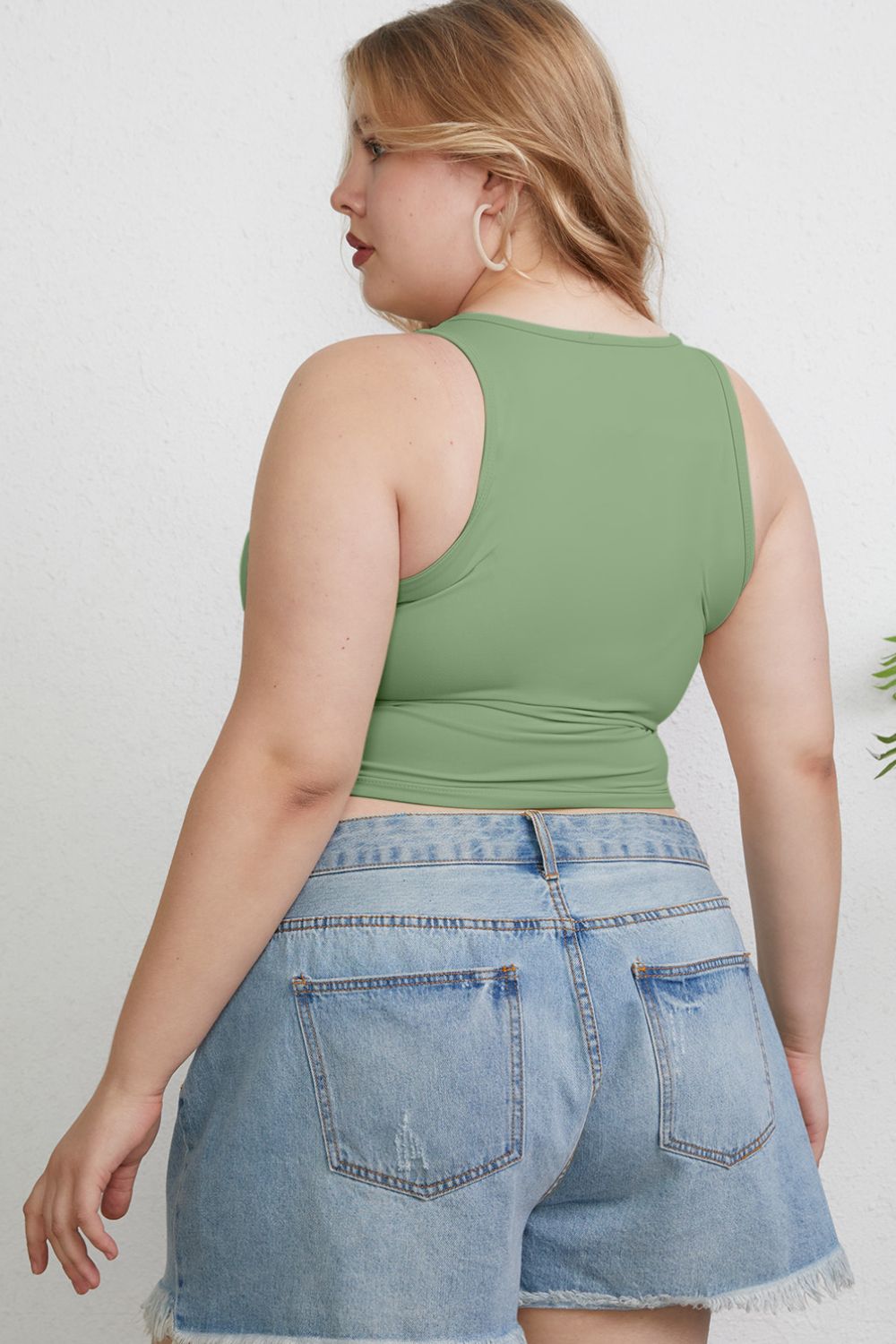 A woman with light hair wears the Basic Bae Full Size Round Neck Slim Tank in black and blue jeans, standing against a light-colored curtain.