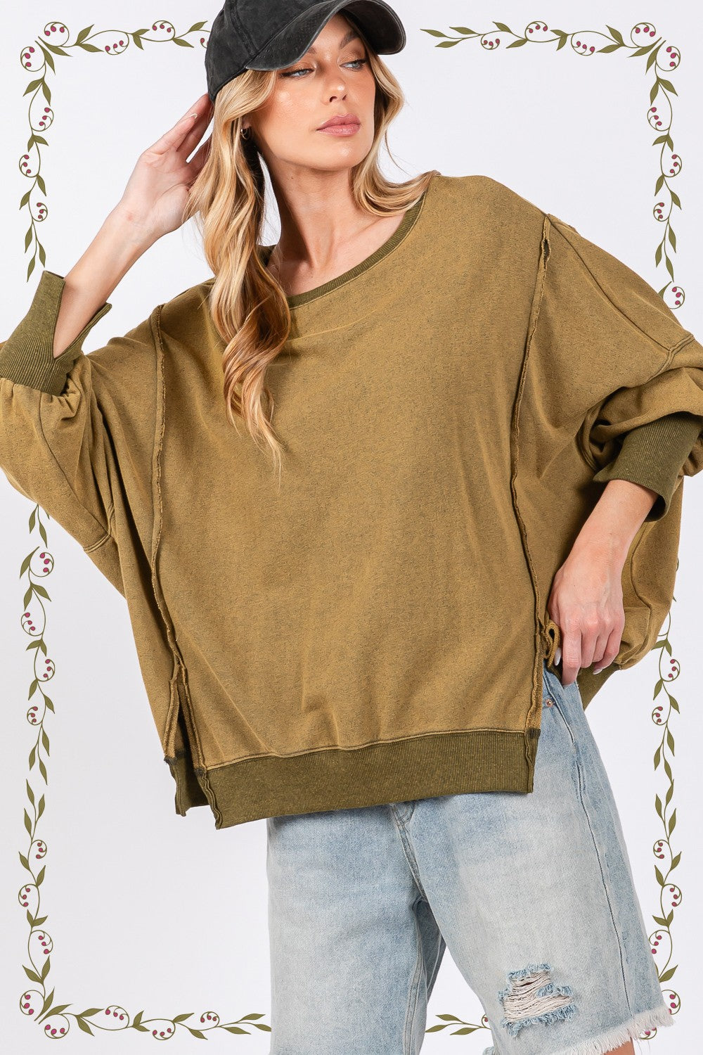 A person wearing a loose, oversized mineral wash SAGE + FIG Side Slit Sweatshirt in olive green, light blue jeans, and a black cap poses against a plain background.