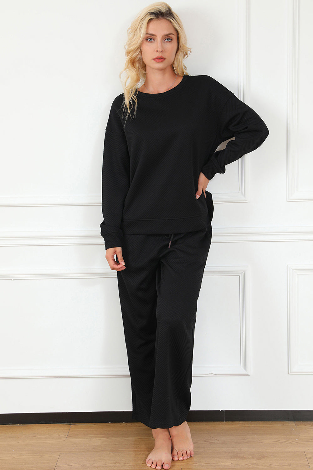 A woman with blonde hair, dressed in the Double Take Full Size Textured Long Sleeve Top and Drawstring Pants Set featuring a stretchy black long-sleeve top and matching wide-leg trousers made of rayon spandex, stands barefoot in a room with white walls and a wooden floor.