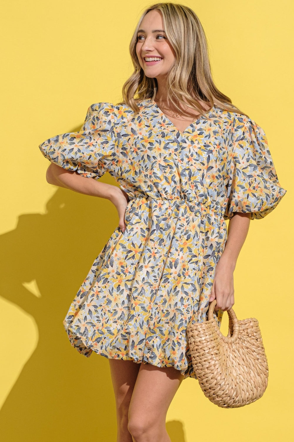 A person wearing the And The Why Full Size Floral Surplice Puff Sleeve Dress stands against a yellow background, holding a woven bag and wearing white sandals.