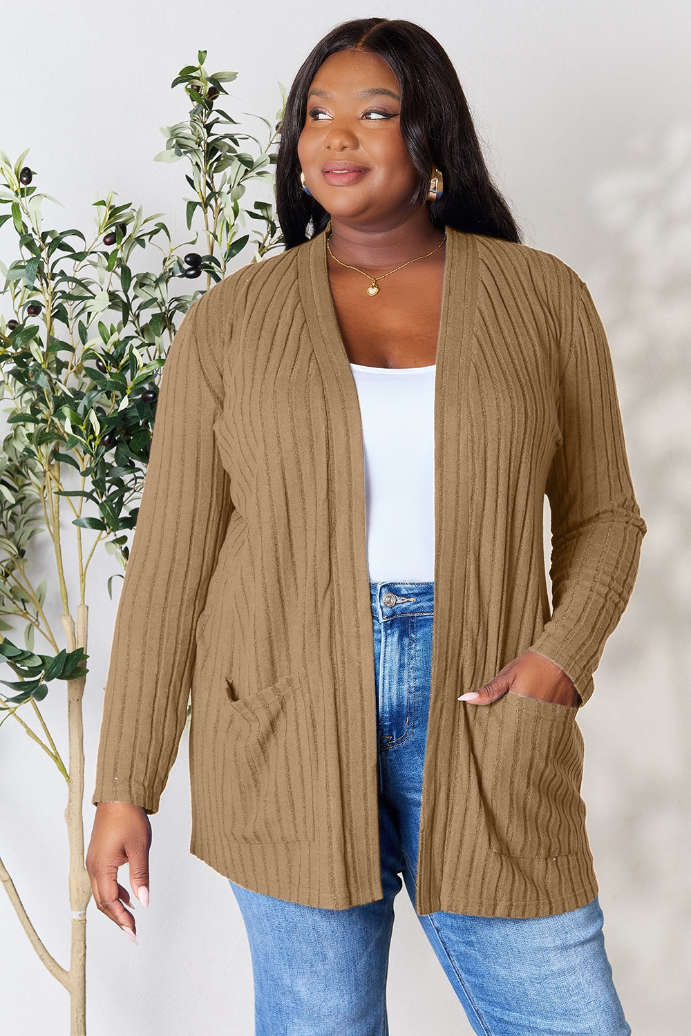 A woman in a Basic Bae Full Size Ribbed Open Front Cardigan with Pockets and jeans, made from stretchy material, stands in front of a plant, smiling slightly and looking to the side.