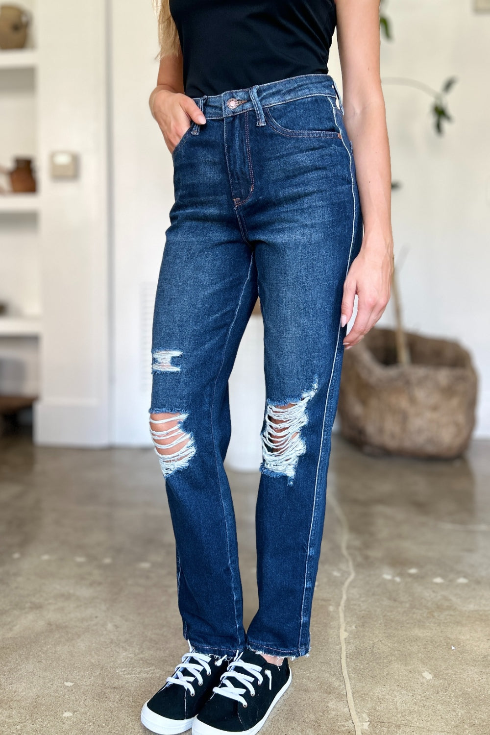 A woman in a black sleeveless top and Judy Blue Full Size High Waist Rigid Magic Heavy Destroy Straight Jeans stands indoors with her hands in her pockets. The background features a plant and shelves adorned with decor items.