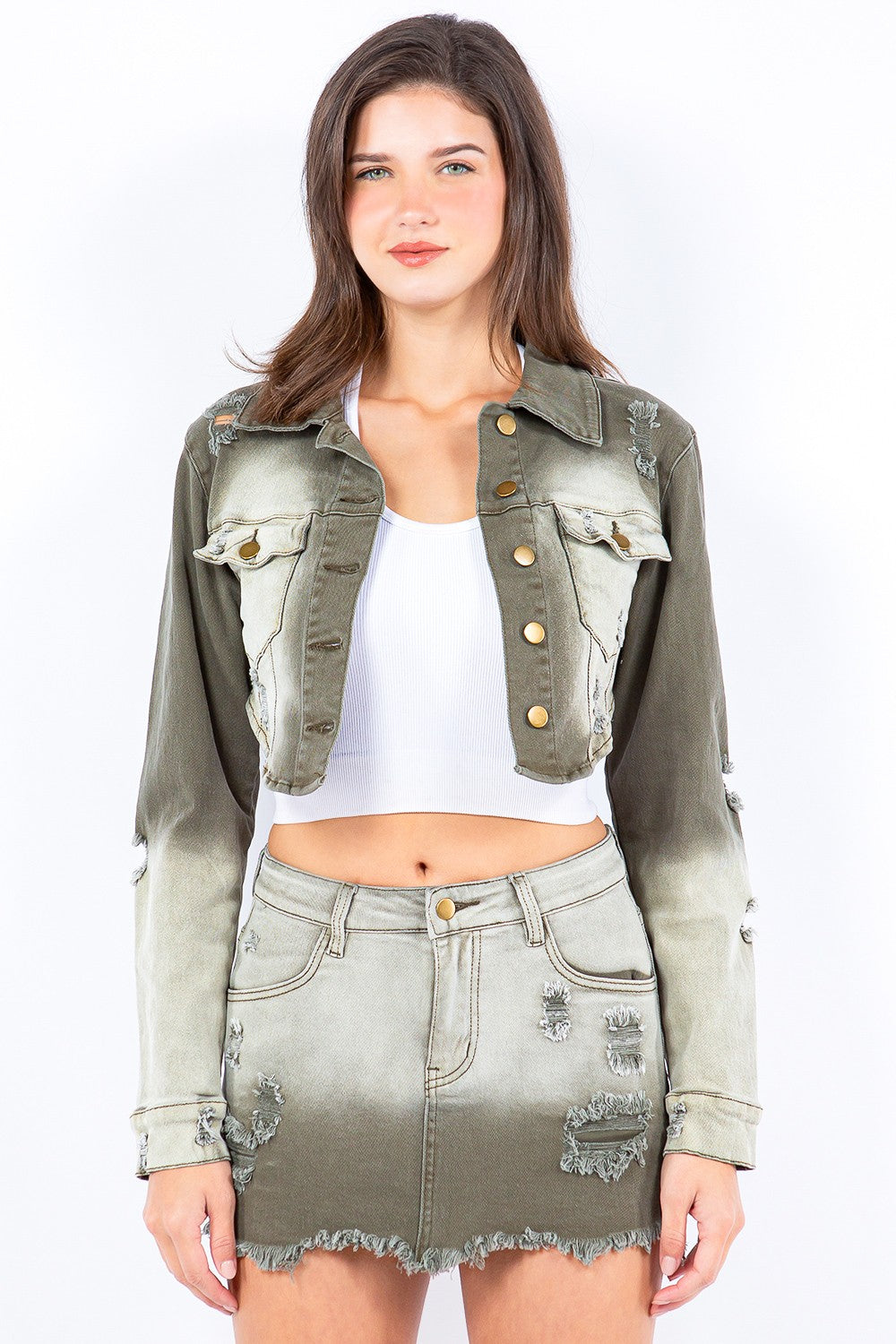 A person stands wearing an American Bazi Distressed Ombre Washed Cropped Denim Jacket in light olive green, paired with a matching skirt featuring a frayed hem and a white cropped tank top.