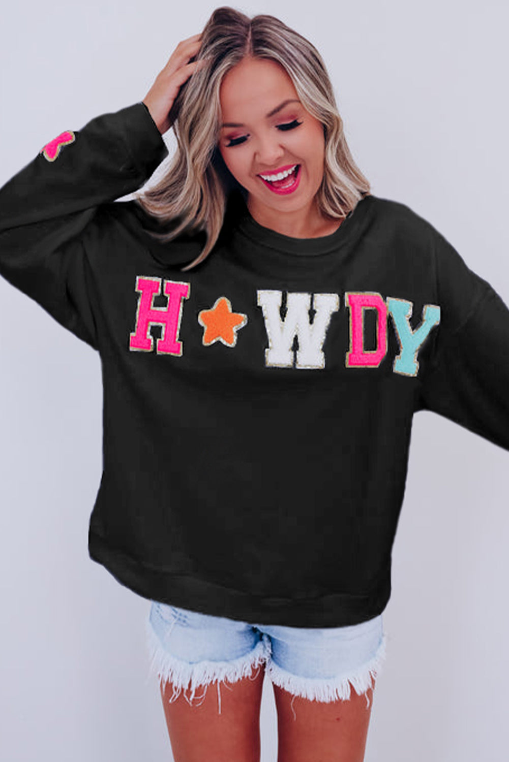 A woman with short blonde hair is wearing the Black Howdy Glitter Chenille Patch Graphic Casual Sweatshirt, featuring colorful star patterns and "WD" letters. She pairs it with light denim shorts and smiles while looking to the side.