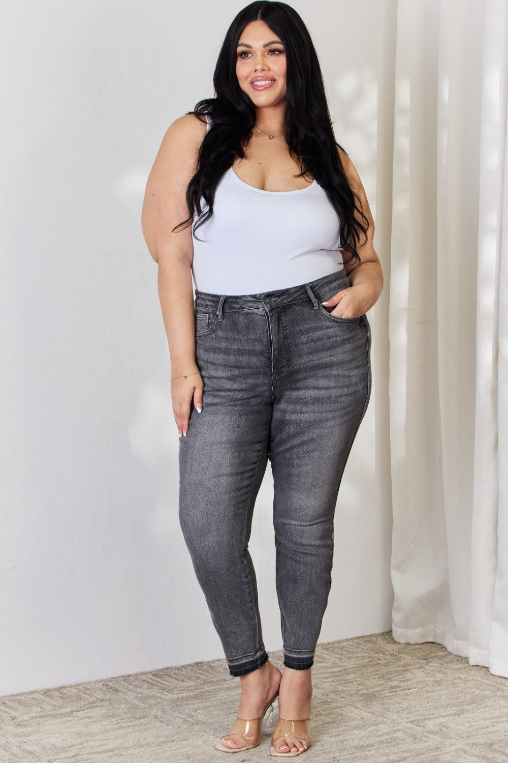 A person wearing the Judy Blue Full Size High Waist Tummy Control Release Hem Skinny Jeans paired with nude heels stands against a light background.