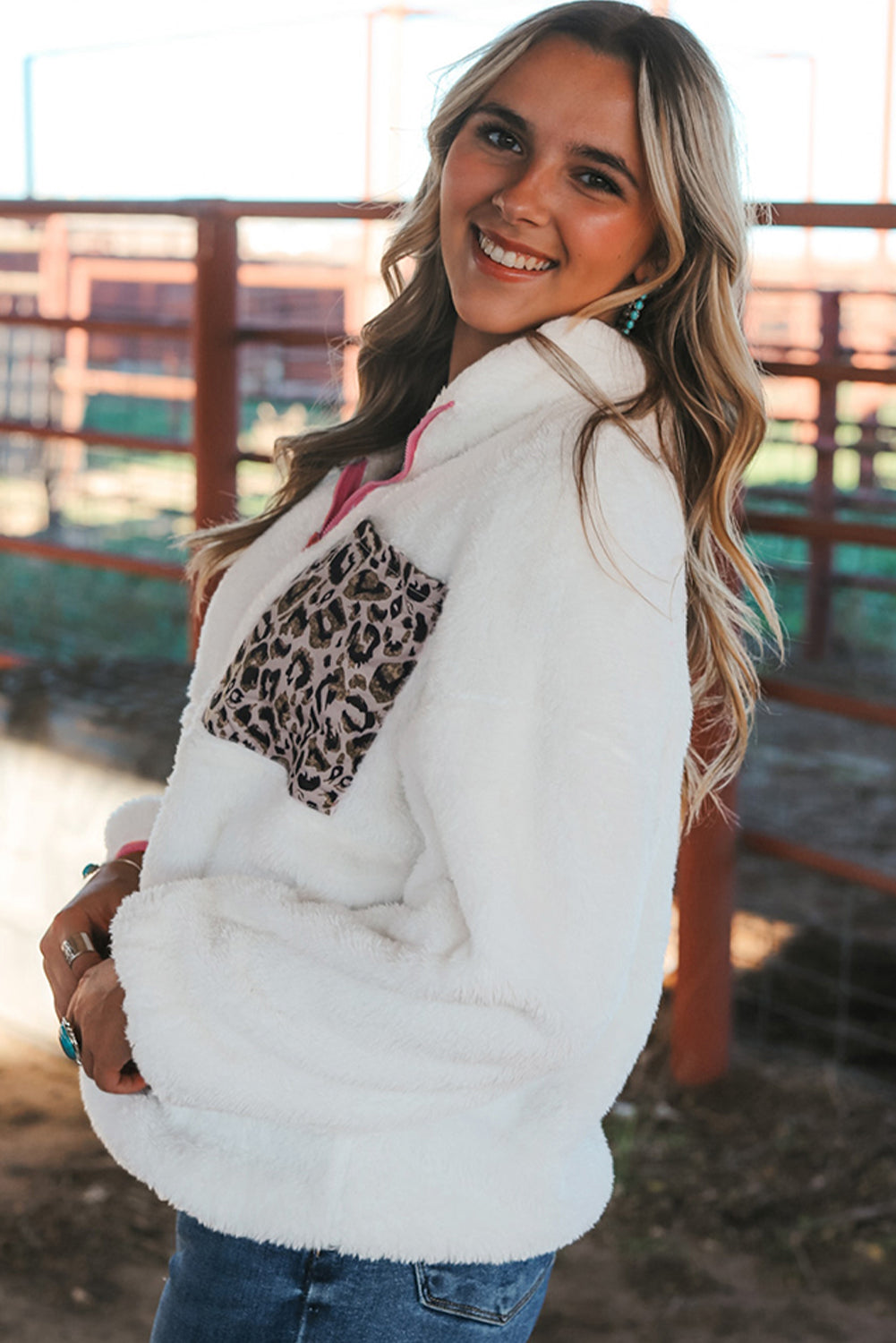 A woman with long, wavy blonde hair is seen from behind, wearing a White Leopard Patch Pocket Half Zipped Fleece Sweatshirt and blue jeans while standing outdoors near red metal fencing.