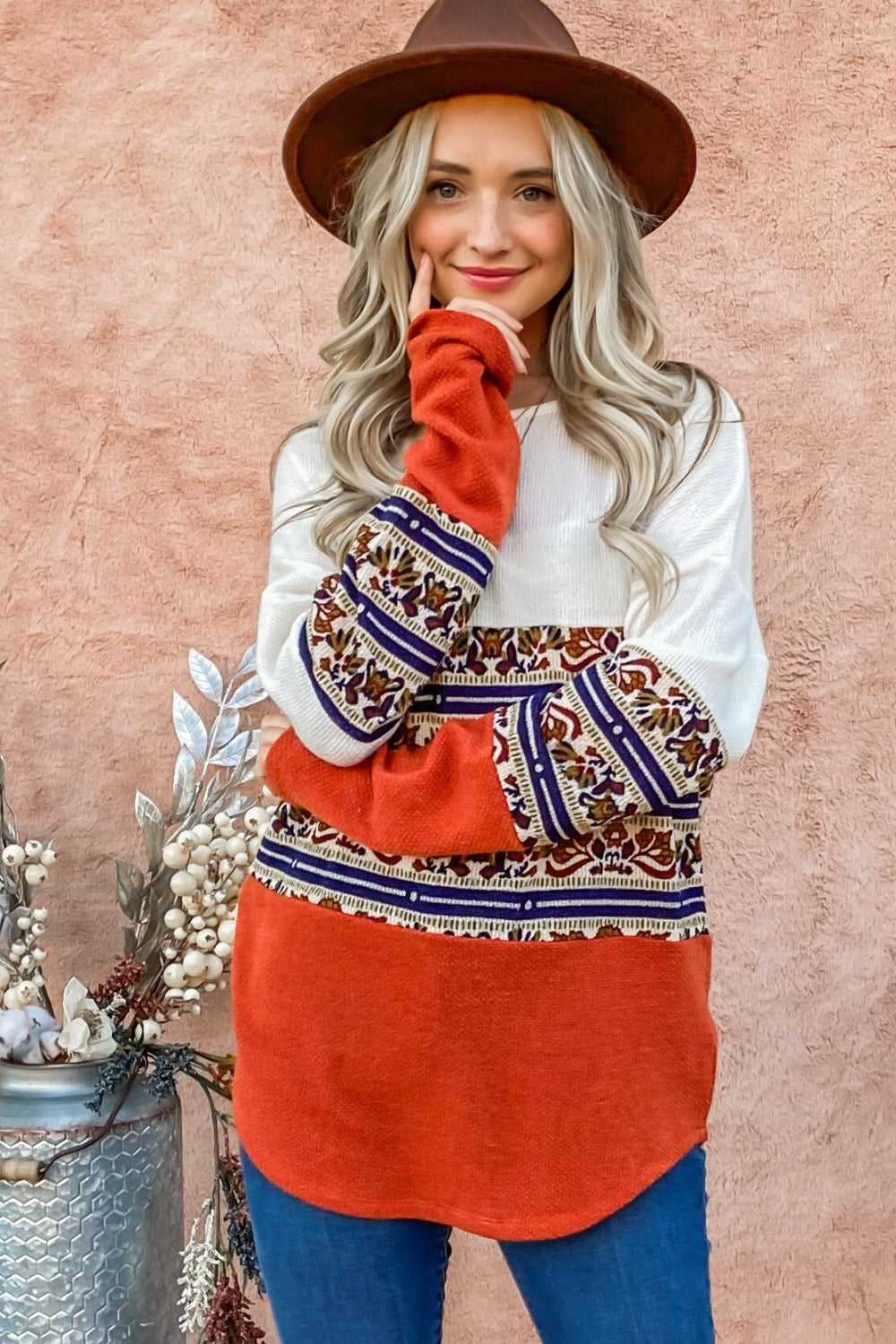 A woman with long blonde hair wearing a brown hat and the And The Why Ethnic Print Color Block Top stands against a peach wall next to a floral arrangement.