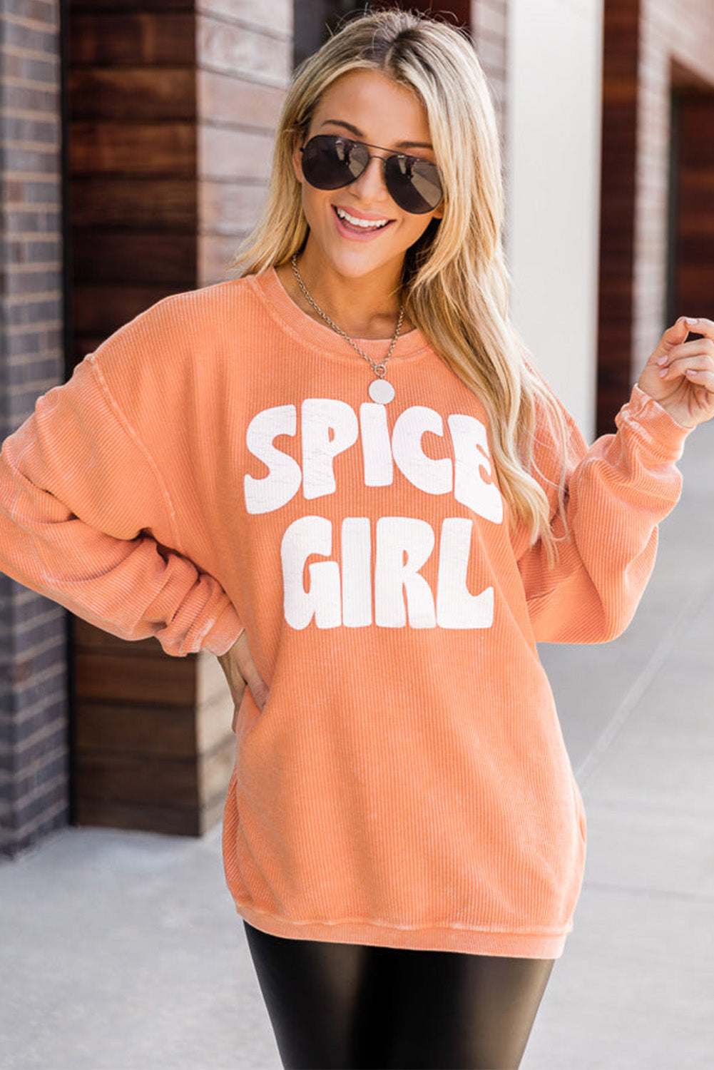 A woman with wavy blonde hair, wearing an Orange Corded SPICY GIRL Graphic Sweatshirt, black leggings, a beige baseball cap, and sunglasses, stands on a street facing away from the camera.