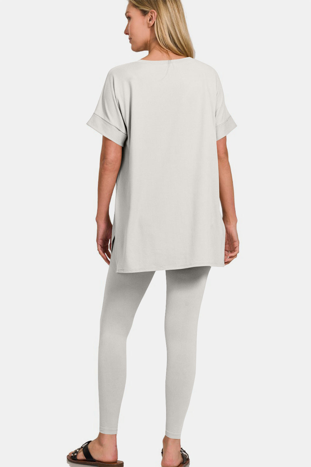 The individual gracefully dons the Zenana V-Neck Rolled Short Sleeve T-Shirt and Leggings Lounge Set in white, standing confidently with one hand on their hip. Paired with sandals, this ensemble captures both comfort and style against a simple backdrop.