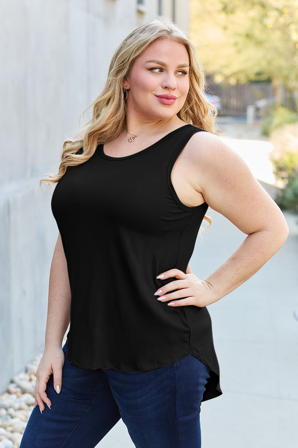 A woman wearing sunglasses, blue jeans, and the Basic Bae Full Size Round Neck Curved Hem Tank is standing in front of a concrete wall, holding her glasses with one hand and carrying a beige shoulder bag. Her basic style complements the slightly stretchy fabric of her outfit, ensuring comfort while maintaining chic simplicity.