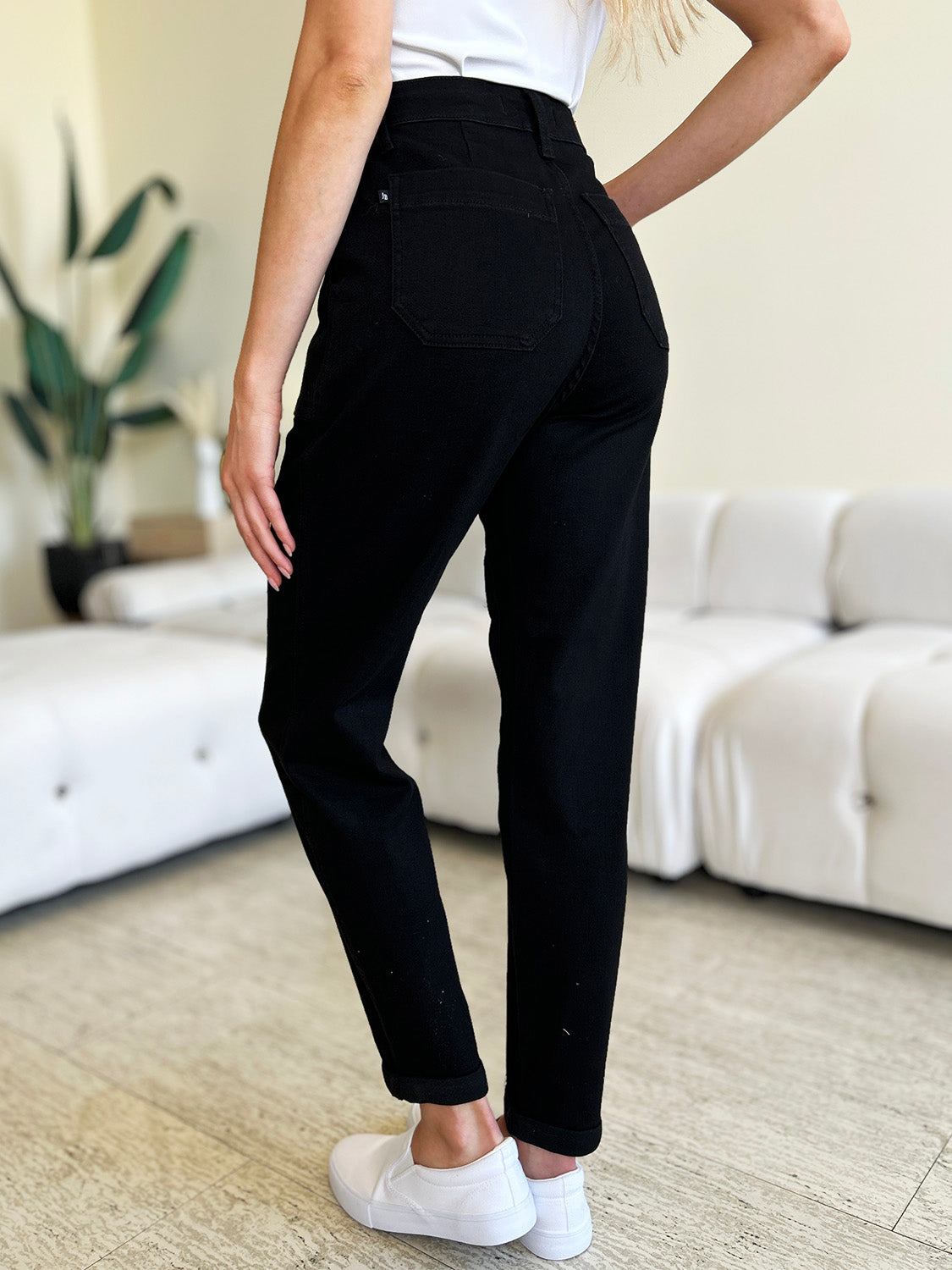 Person standing indoors, wearing a white top, black Judy Blue Full Size High Waist Double Roll Cuff Jeans, and white slip-on shoes, next to white couches and a plant in the background.