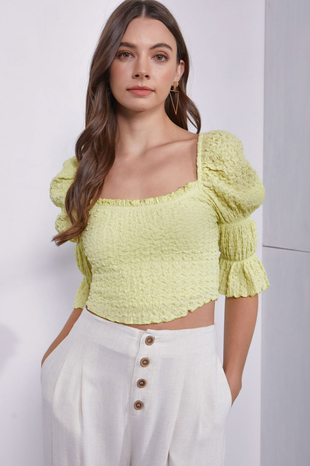 A woman with long brown hair wearing a trendy and stylish "Mustard Seed Crinkle Texture Puff Sleeve Crop Top" and high-waisted white pants with buttons stands against a plain background.