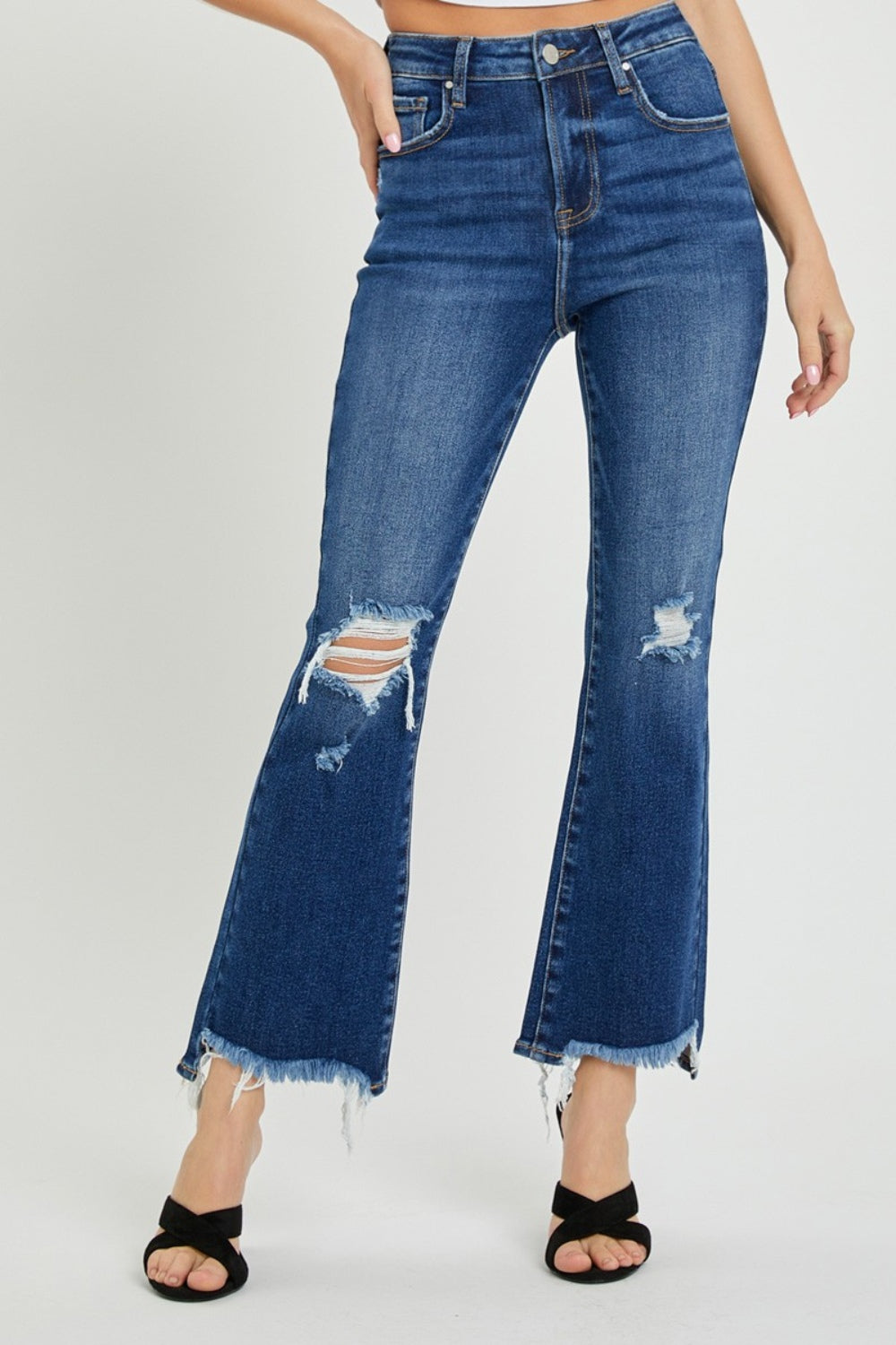 A person wearing RISEN High Rise Distressed Crop Flare Jeans with frayed hems stands with their right hand on their left hip, paired with black open-toed sandals.