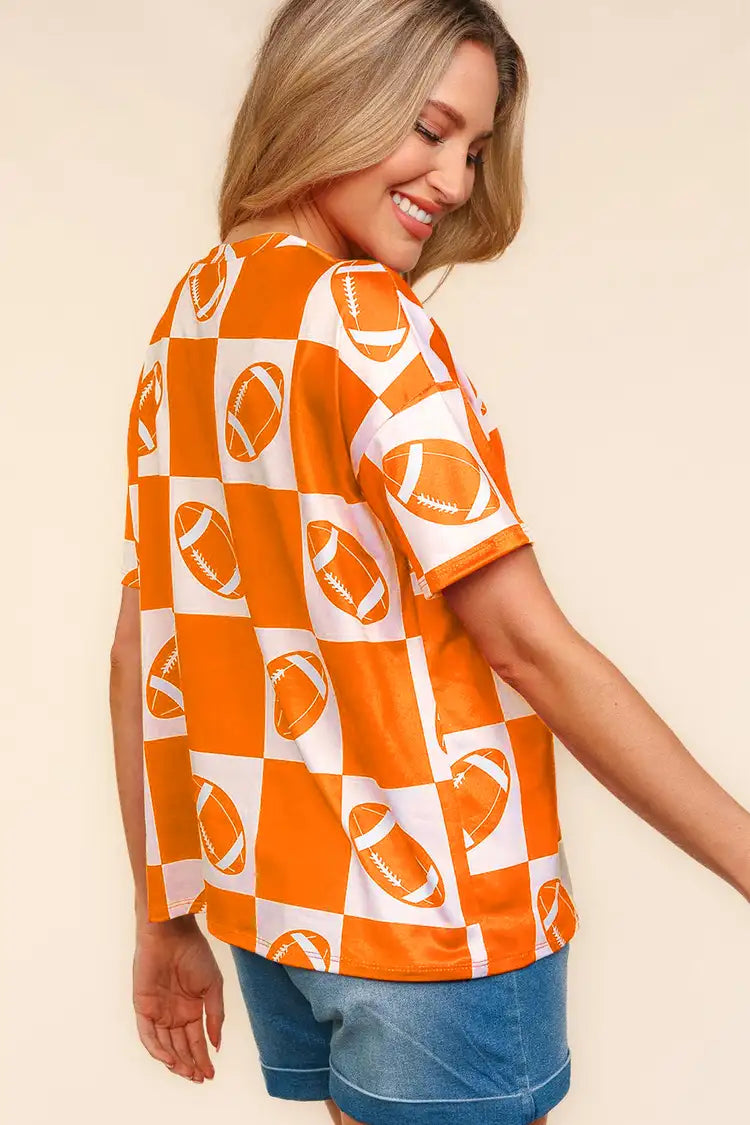 A woman wears a Haptics Football Checkered Print Short Sleeve T-Shirt in orange and white, standing against a plain background, and pairs it perfectly with denim shorts for a casual sporty look.