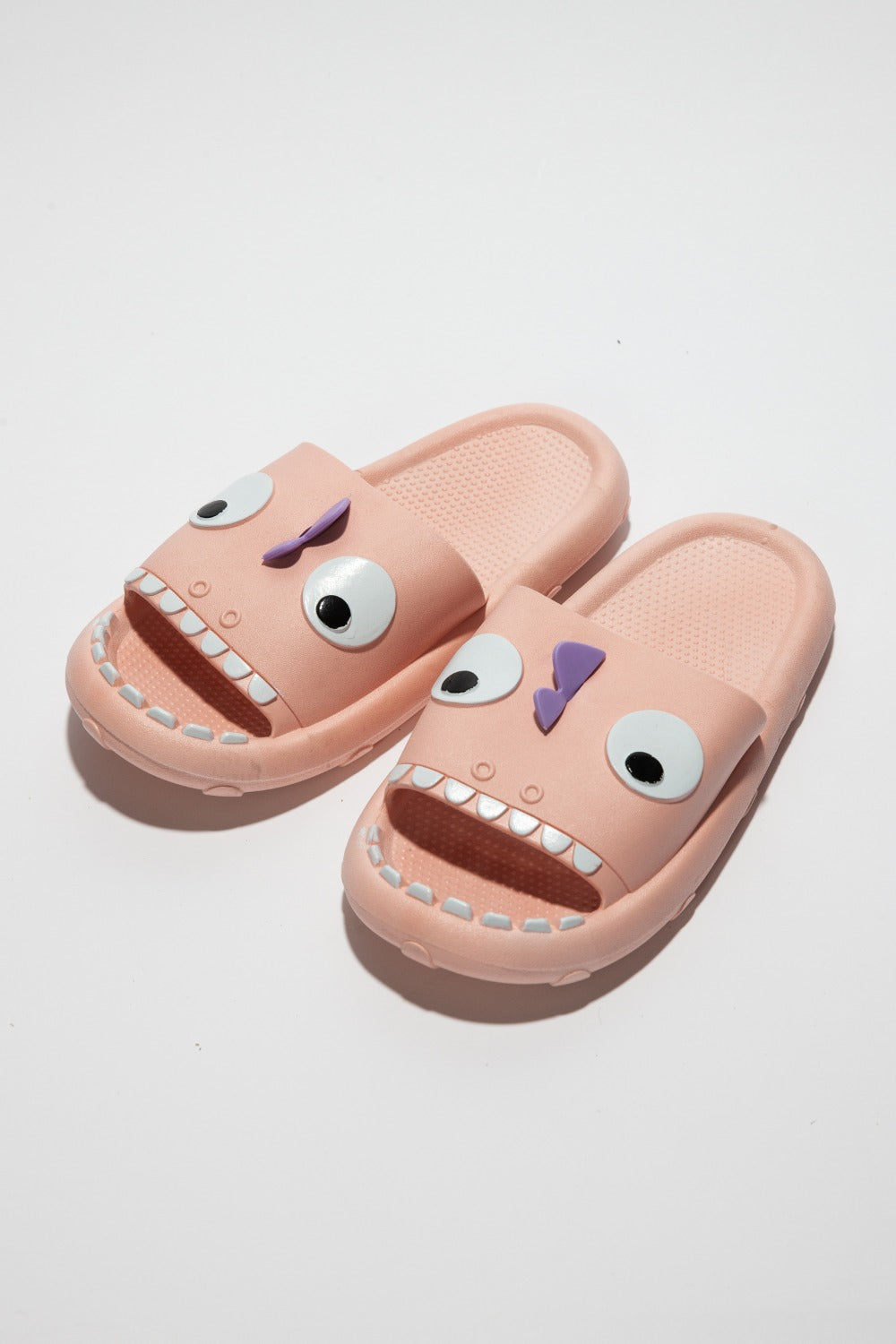 The NOOK JOI Monster Pillow Cloud Slides Non-Slip Slipper features cartoonish monster faces with large eyes, a purple nose, and an open mouth with teeth. Made of durable EVA material, these fun pink cloud slippers also boast an anti-slip design for added safety.