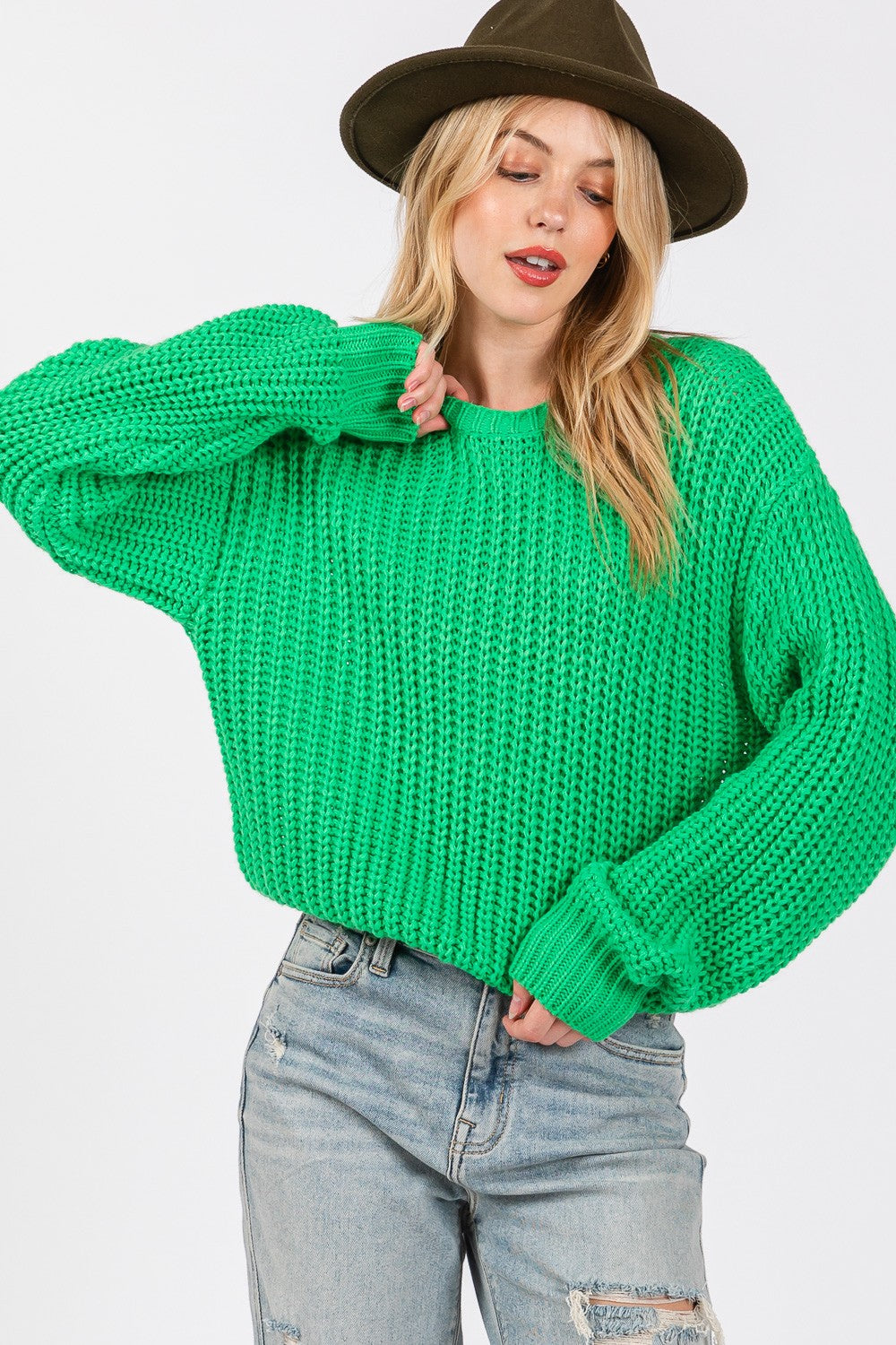 A person wearing a SAGE + FIG round neck drop shoulder sweater, ripped light blue jeans, and a green hat poses against a white background.