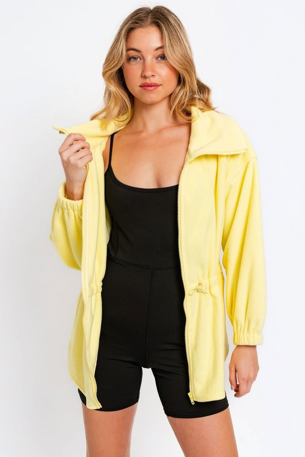 A woman wearing a black outfit and a yellow Le Lis Zip Up Waist Drawstring Soft Fleece Jacket stands against a plain white background.