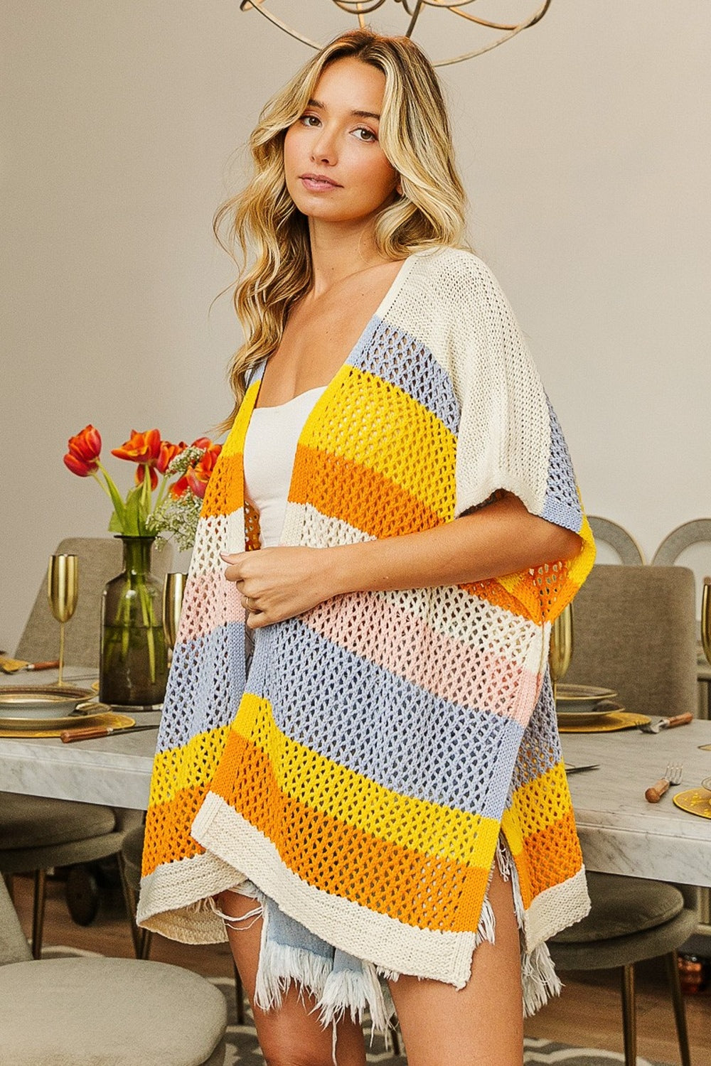 A woman in a brightly colored BiBi Openwork Striped Open Front Knit Cardigan and shorts stands in a dining room with a set table and a vase of flowers, showcasing this versatile layering piece.