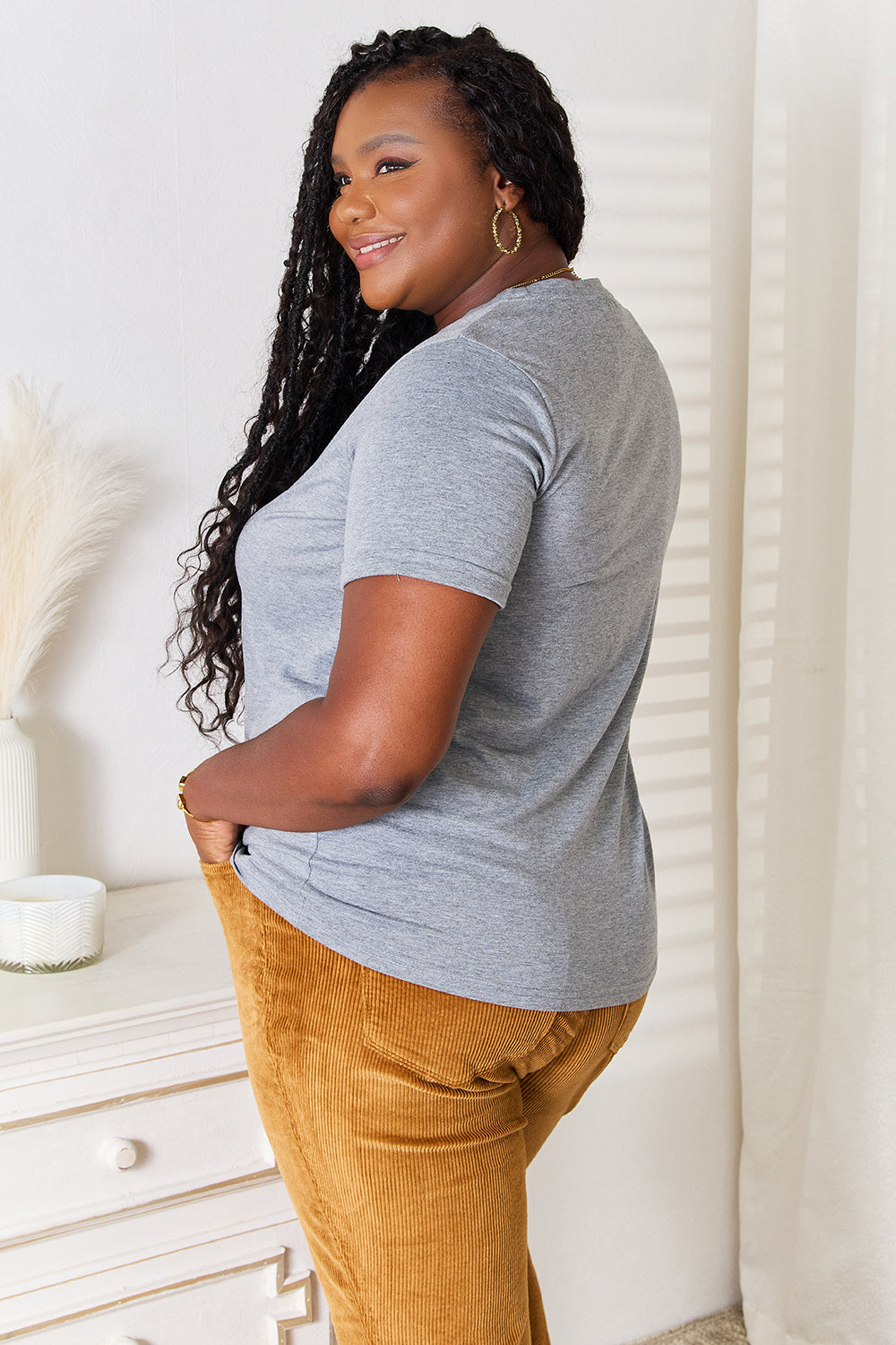 A woman with long dark hair is wearing a Simply Love US Flag Graphic Cuffed Sleeve T-Shirt and brown pants as she stands indoors against a light-colored background.