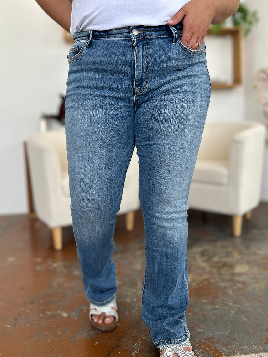 Person wearing Judy Blue Full Size Mid-Rise Waist Straight Jeans and a black top, standing in a room with white chairs, potted plants, and wooden shelves. Only the lower half of the body and part of the arms are visible.