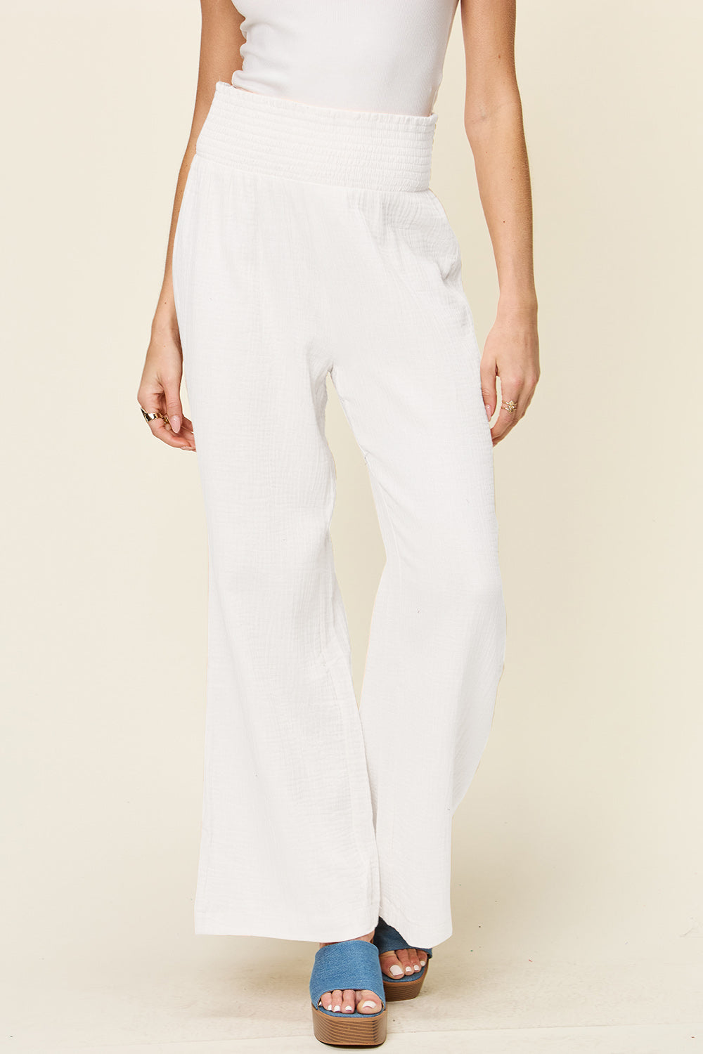 A person wearing Double Take Full Size Texture Smocked Waist Wide Leg Pants in black and a white 100% cotton top is standing with one hand on their hip. They are also wearing blue open-toe sandals.