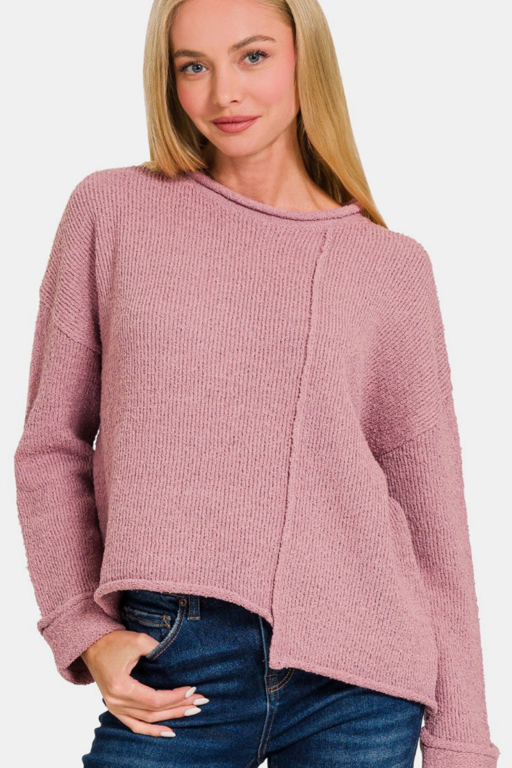 A person with long blonde hair is wearing a Zenana Asymmetric Hem Drop Shoulder Sweater in light pink and blue jeans, standing against a plain white background.