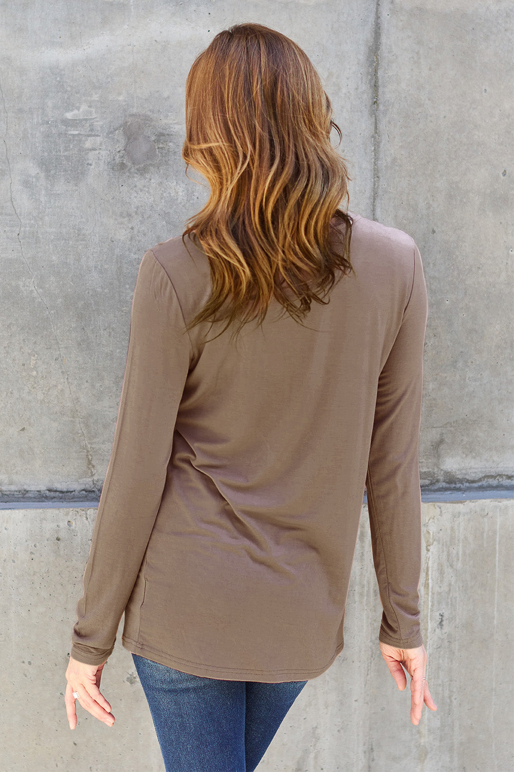 A woman with shoulder-length hair wearing the Basic Bae Full Size Round Neck Long Sleeve Top and blue jeans stands in front of a concrete wall, holding a beige handbag over her shoulder. Her outfit exemplifies basic style, with the top made from 100% polyester and recommended for machine wash cold.