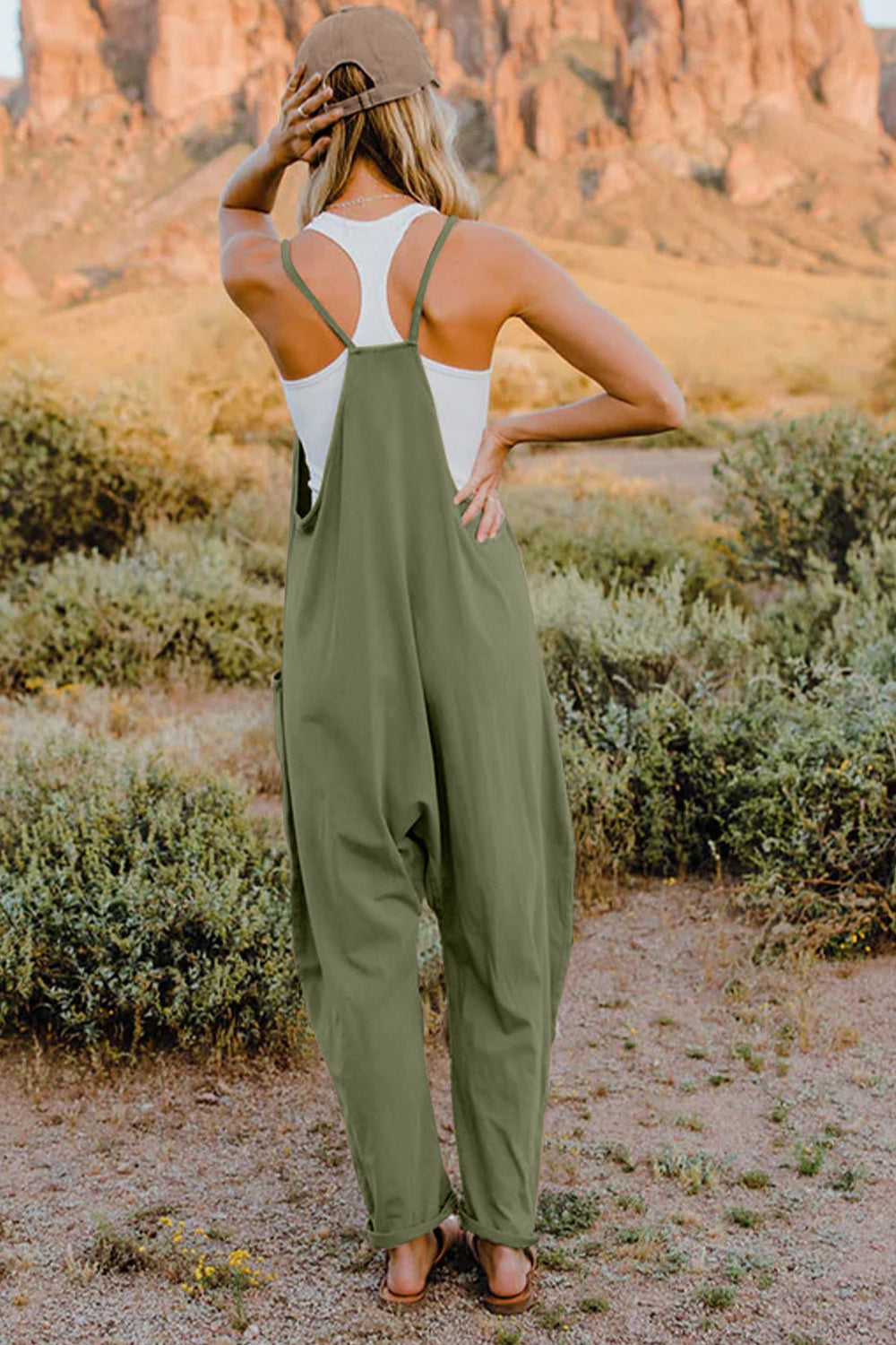 Person wearing a Double Take Full Size V-Neck Sleeveless Jumpsuit with Pockets and sandals, walking outdoors on a dirt trail with bushes in the background.