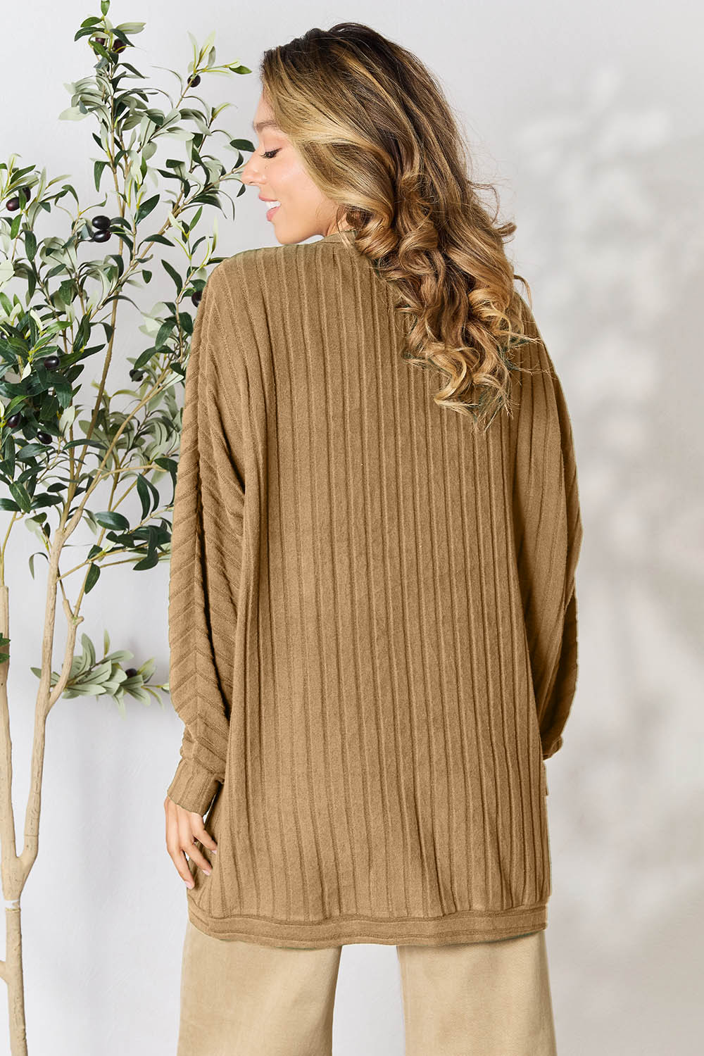 A person with wavy hair, wearing the Basic Bae Full Size Ribbed Cocoon Cardigan over a white top and beige pants, stands in front of a light-colored background with a leafy plant, showcasing an effortlessly basic style.