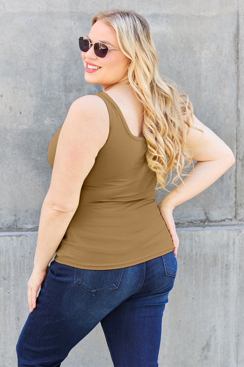 A woman with brown hair, wearing sunglasses, blue jeans, and the Basic Bae Full Size Square Neck Wide Strap Tank in black stands against a concrete wall, showcasing a basic style.