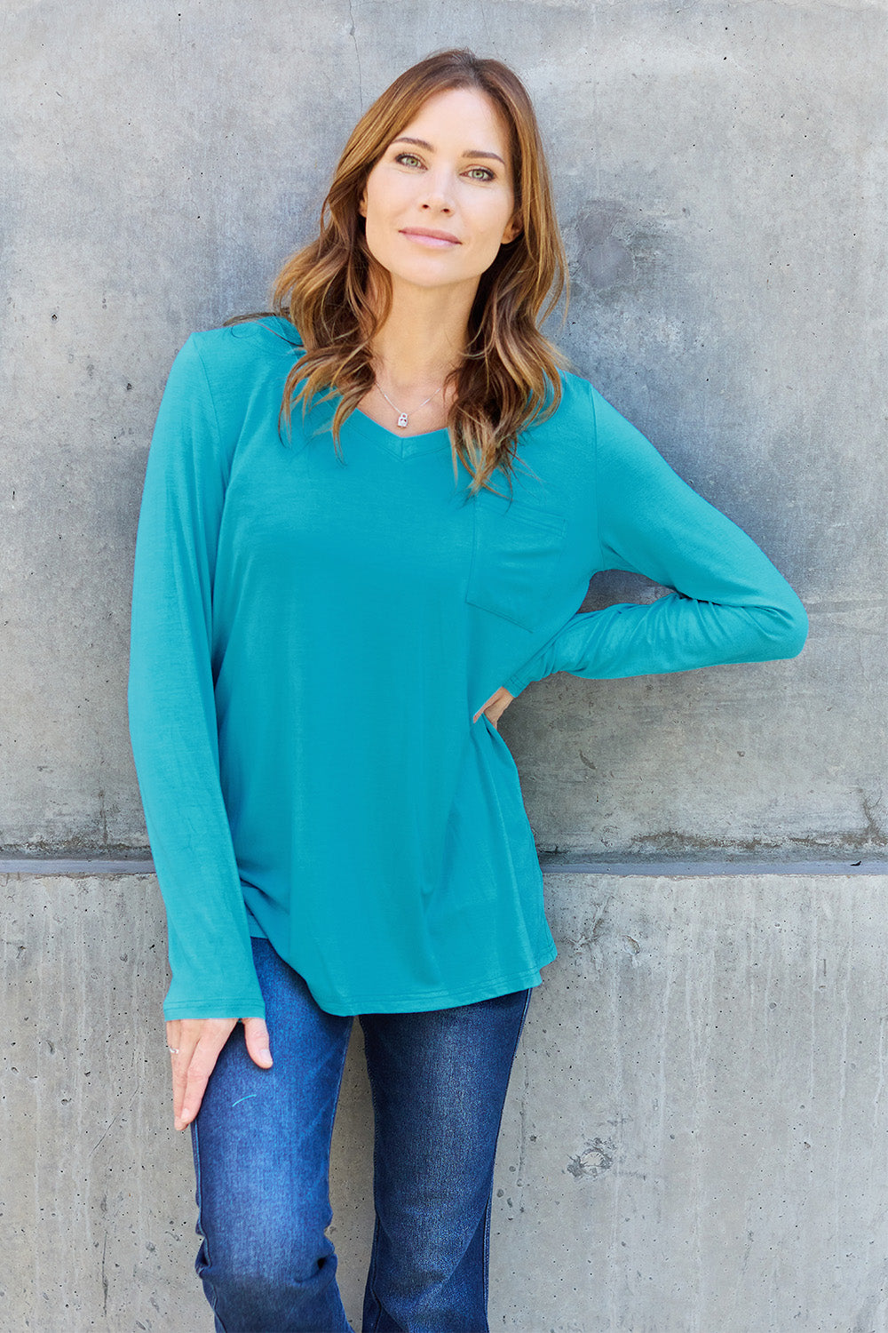 A woman with light brown hair stands against a concrete wall, wearing the Basic Bae Full Size V-Neck Long Sleeve Top in olive green and blue jeans, with a white backpack on her shoulder. Her outfit exudes basic style and is made from polyester, ensuring it’s both comfortable and suitable for machine wash cold. She is looking at the camera.