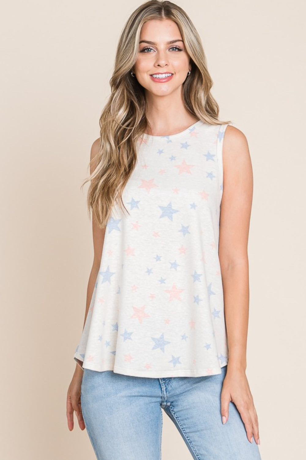 A woman with long, wavy hair is standing and smiling against a beige background, dressed in a casual wear ensemble featuring the BOMBOM Star Print Round Neck Tank with blue and pink patterns, paired with light blue jeans.