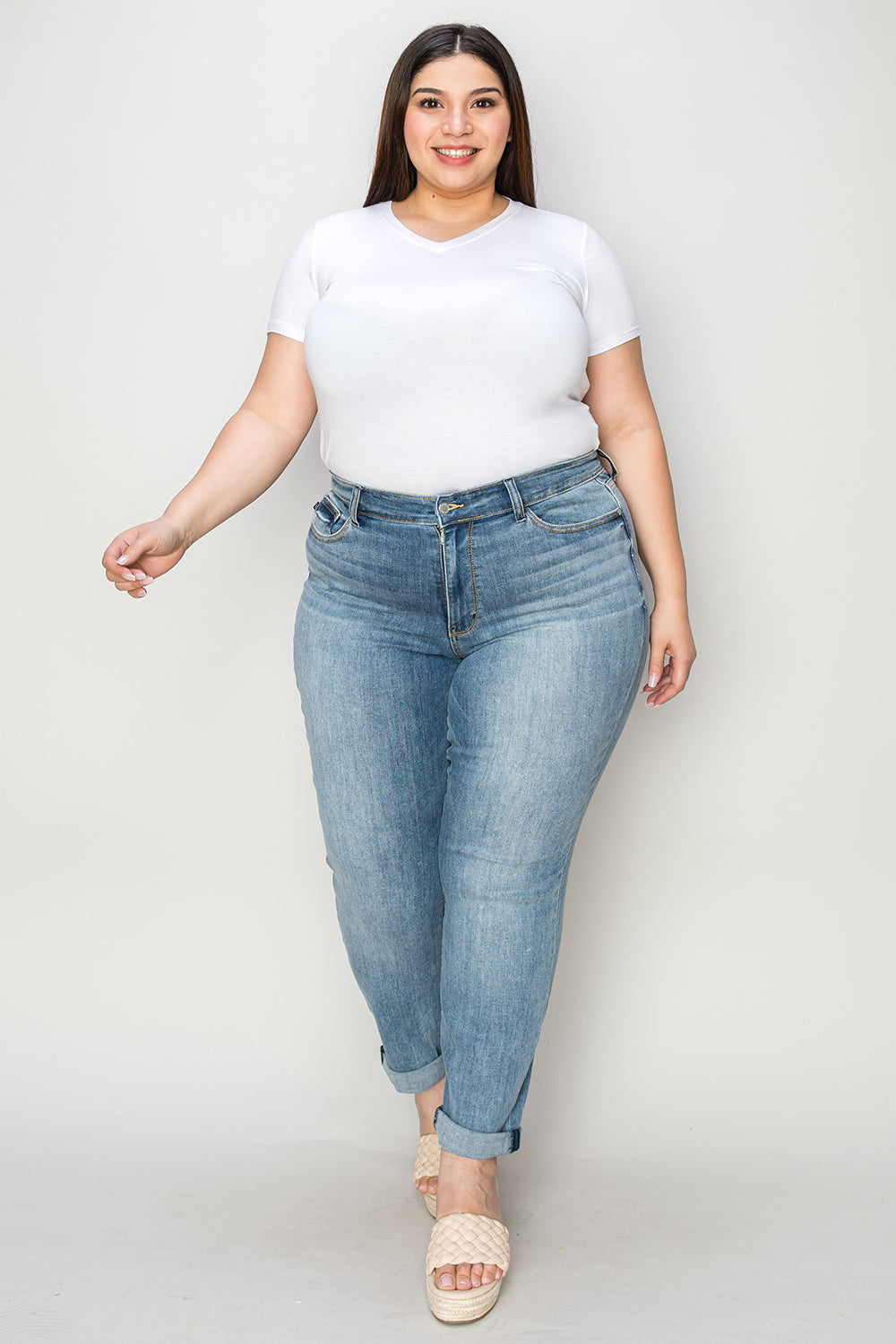 A person wearing a white shirt, Judy Blue Full Size Cuffed Hem Slim Jeans, and black wedge-heeled sandals is standing in a room with beige chairs and plants in the background.