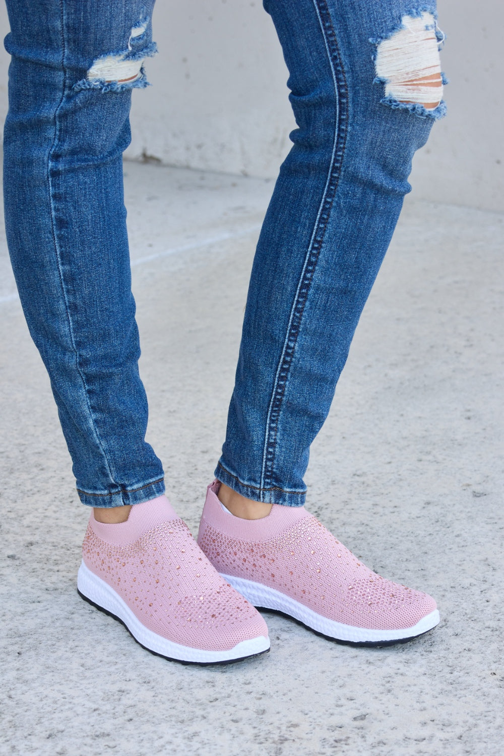 Person wearing Forever Link Rhinestone Mesh Breathable Sneakers in light pink and ripped blue jeans, standing on a concrete surface.