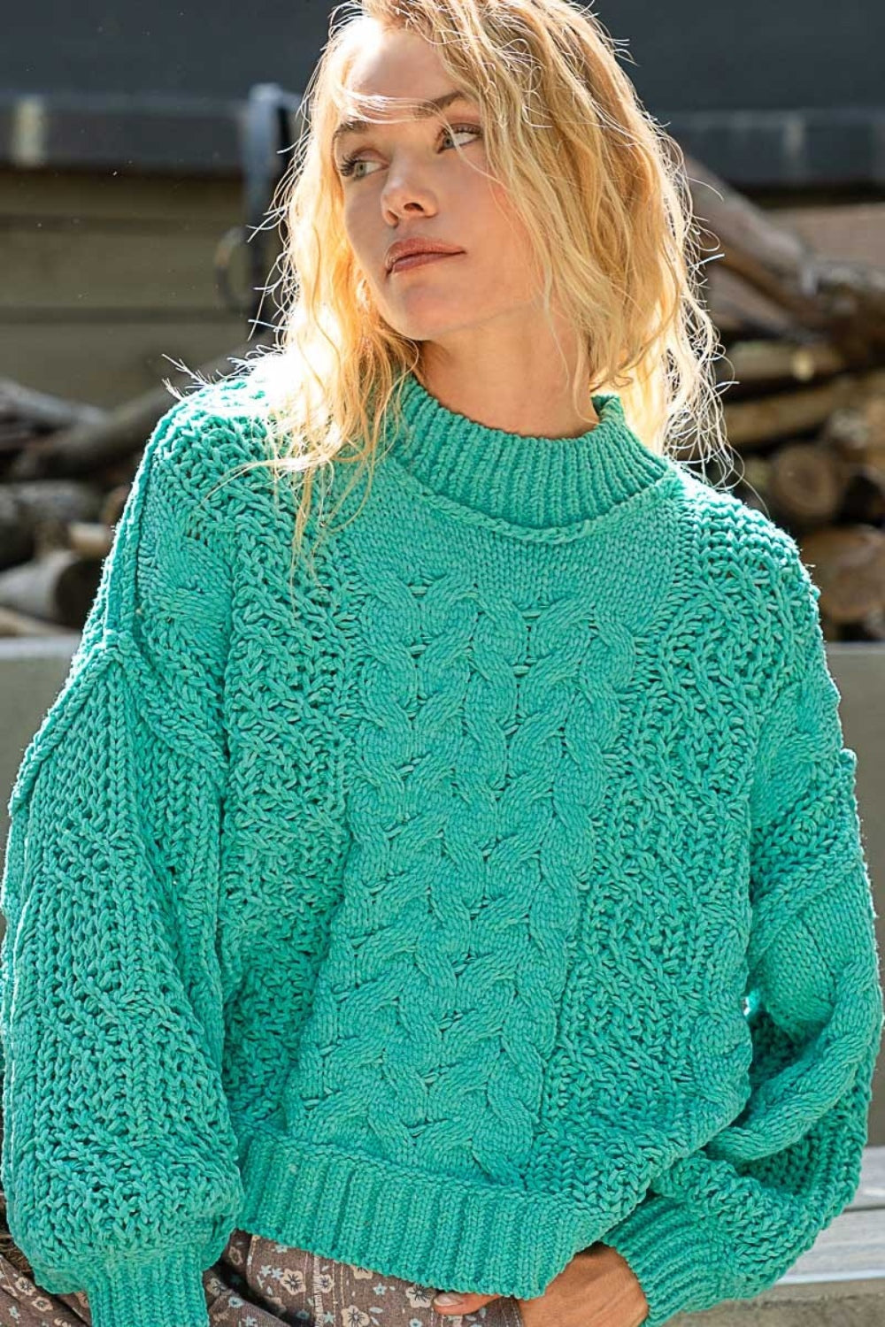 A person in a turquoise POL Mock Neck Cable Knit Sweater with a textured pattern is sitting outdoors.