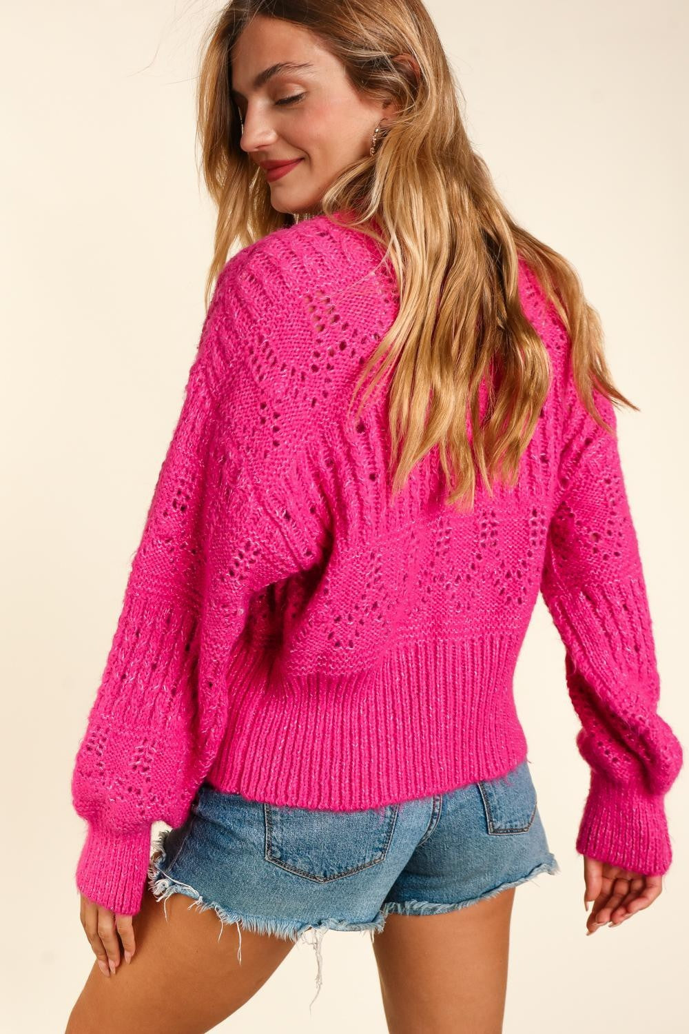 A person with long hair wears the trendy, bright pink Haptics Full Size Button Down Crop Long Sleeve Cardigan over a white top and denim shorts. They hold one side of the cardigan's collar and smile at the camera, showcasing their stylish and versatile layering.