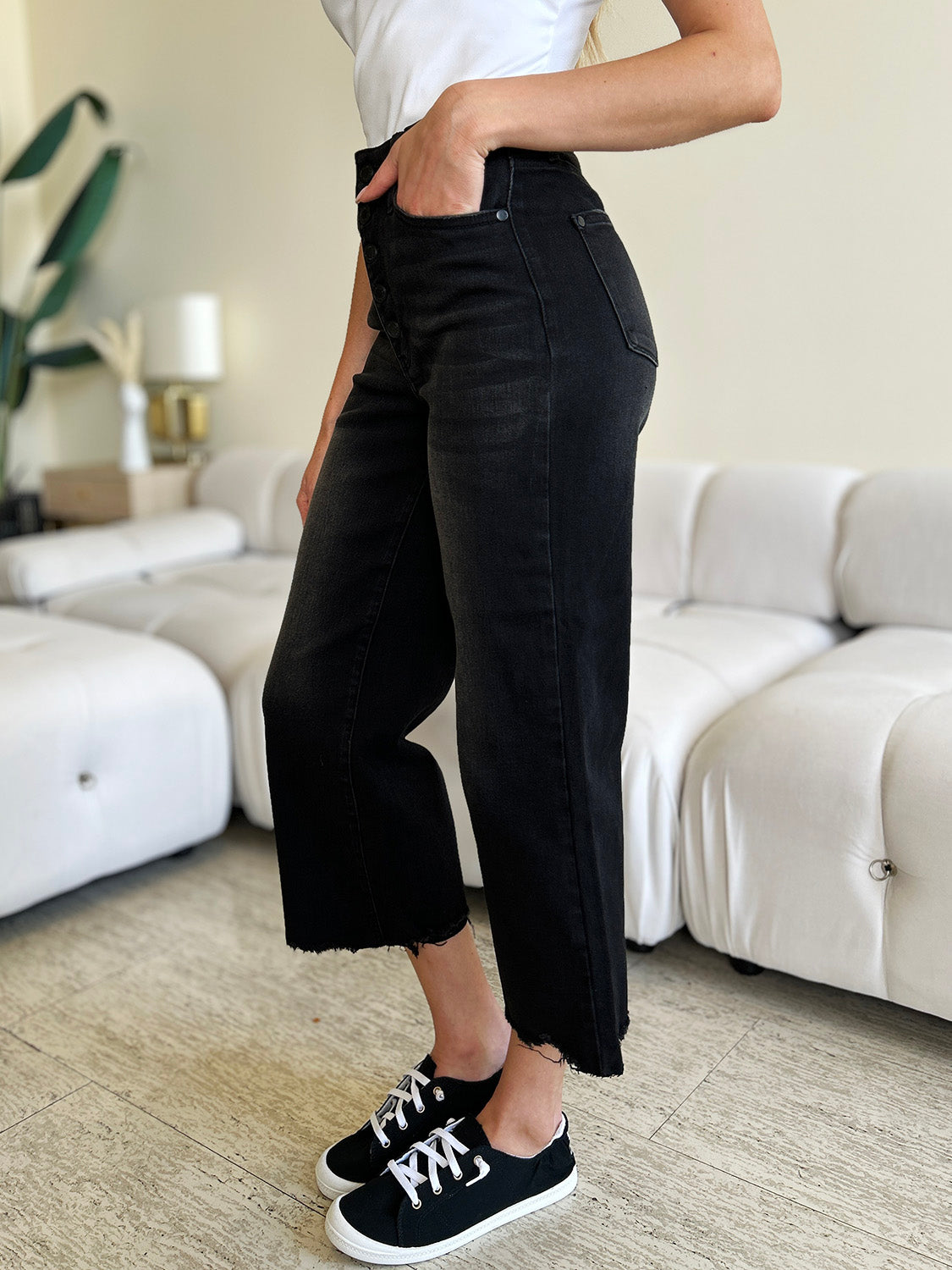 Person wearing Judy Blue Full Size High Waist Button Fly Jeans and black sneakers, standing indoors. Hands are in pockets, and the background includes a white couch and a plant. These high-waisted jeans make the outfit sleek and stylish.