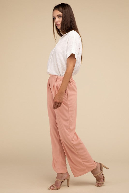 A woman stands against a beige background, highlighting her chic ensemble with a tailored fit. She wears a white short-sleeve shirt paired with versatile pink Waffle Trouser Pants, hands casually in pockets, complemented by stylish strappy sandals.